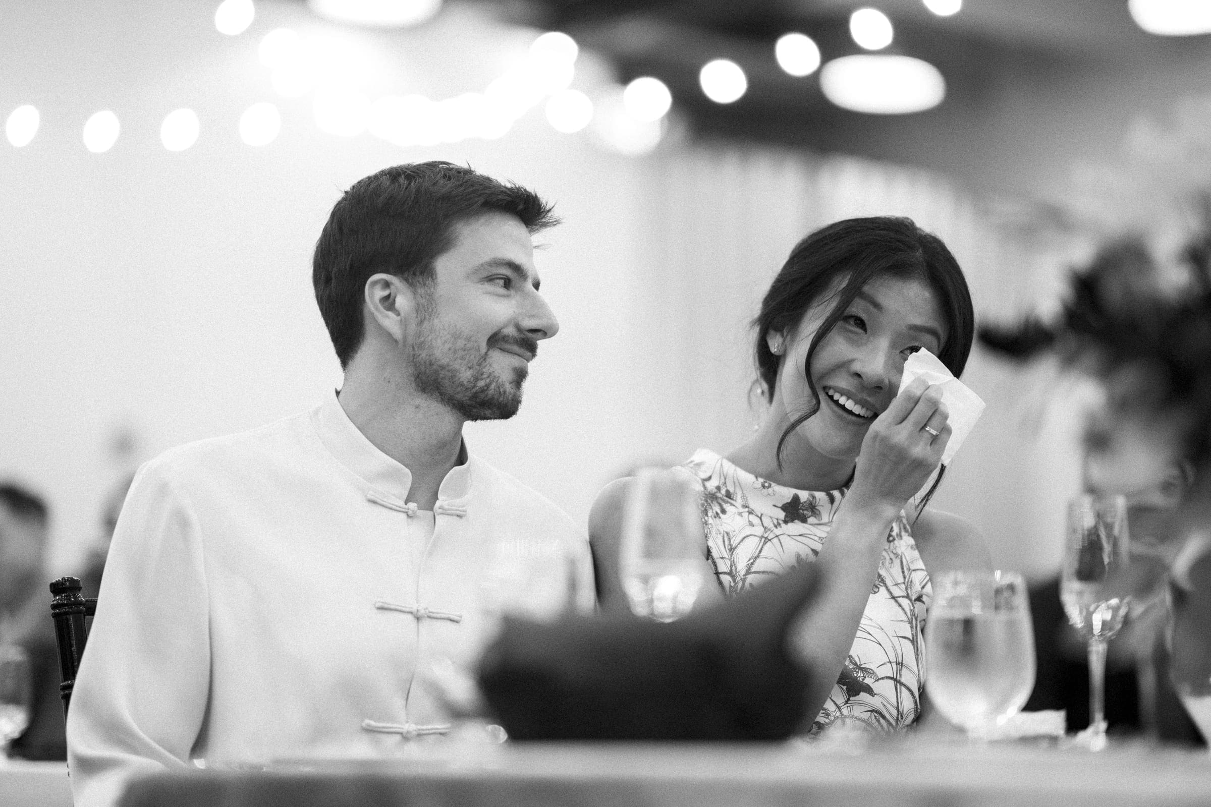 A black and white photo captures a couple seated at a table during their market hall wedding celebration. The man, in a traditional buttoned shirt, smiles warmly while the woman wipes away tears of joy with a tissue, surrounded by drinks and blurred decorations.