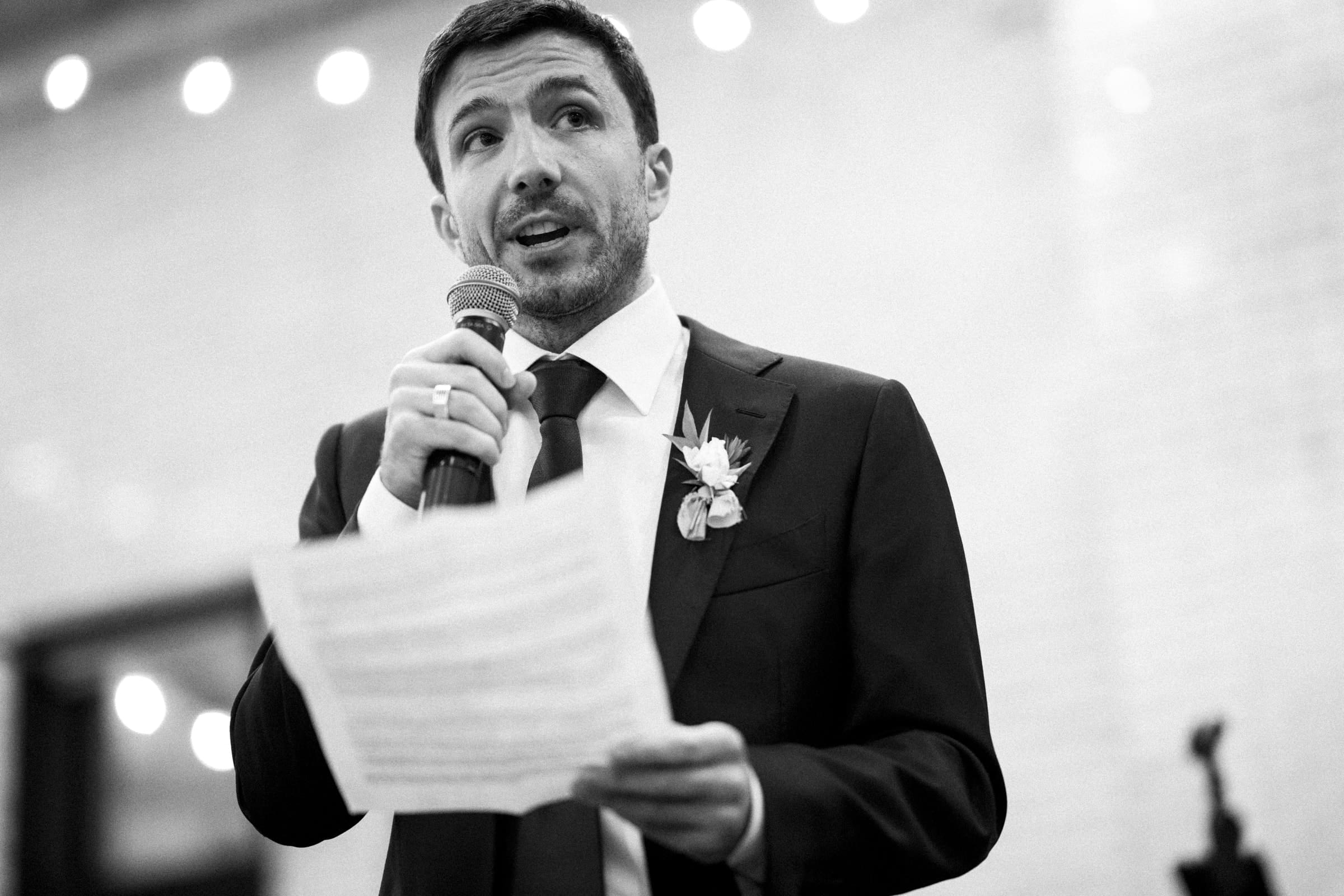 A man in a suit and tie stands holding a microphone and a piece of paper, speaking at what appears to be a sophisticated market hall wedding. The scene is black and white, with string lights in the blurred background, creating a formal and elegant atmosphere.