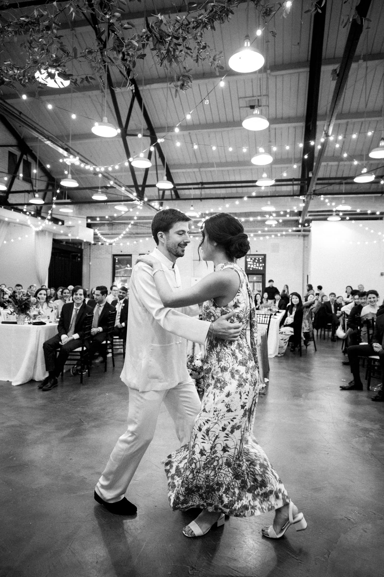 A black and white photo captures a couple dancing at an indoor market hall wedding. The man is in a light suit, and the woman wears a floral dress. Guests are seated around them at tables, while strings of lights elegantly decorate the ceiling.