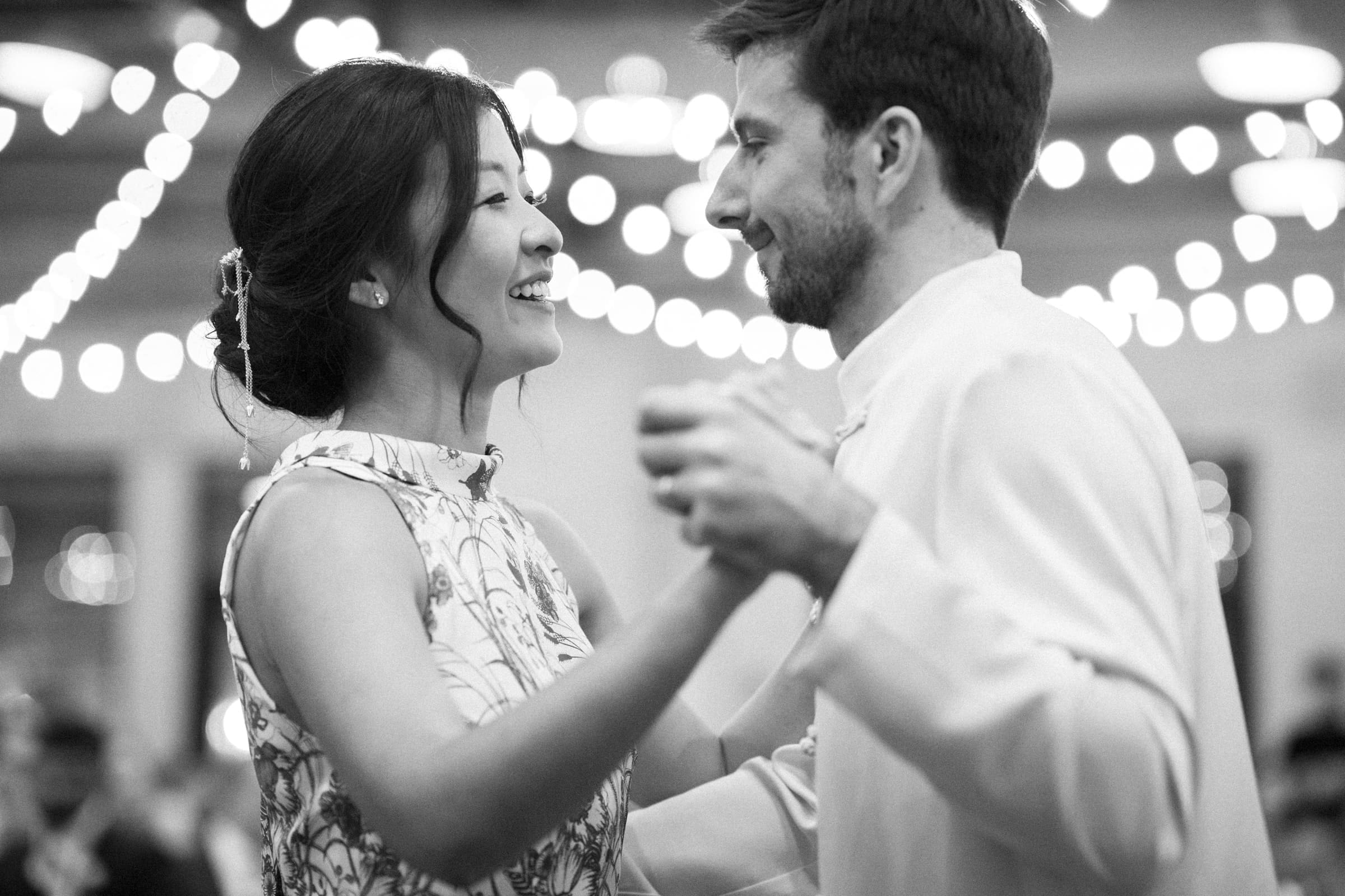 A black and white photo captures a couple elegantly dancing at a market hall wedding. The woman wears a floral dress with her hair styled in an updo, and the man is in a light-colored suit. String lights hang above, adding to the festive atmosphere.