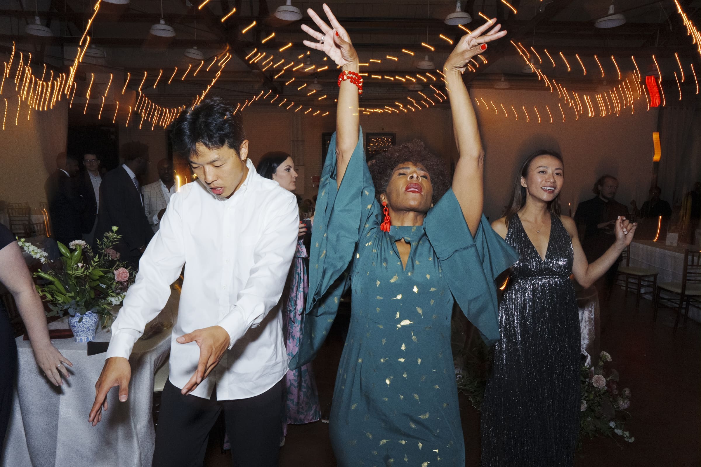 Three people are dancing energetically at a festive market hall wedding under string lights. The person in the center has their hands raised, expressing joy. The atmosphere is lively, with other guests and decorations visible in the background.