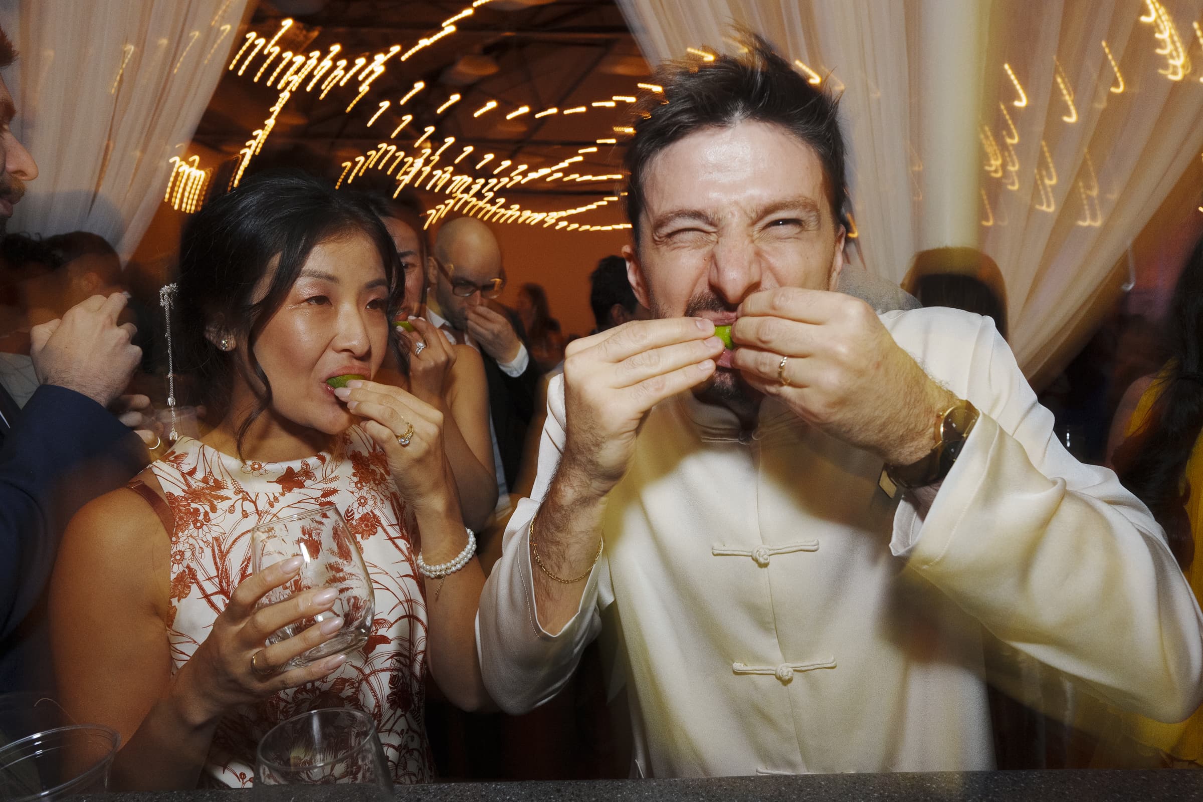A woman and a man are joyfully biting into lime wedges at a lively market hall wedding. The background features string lights and draped curtains, creating a festive atmosphere. Several other people are visible, enjoying themselves in the dim lighting.