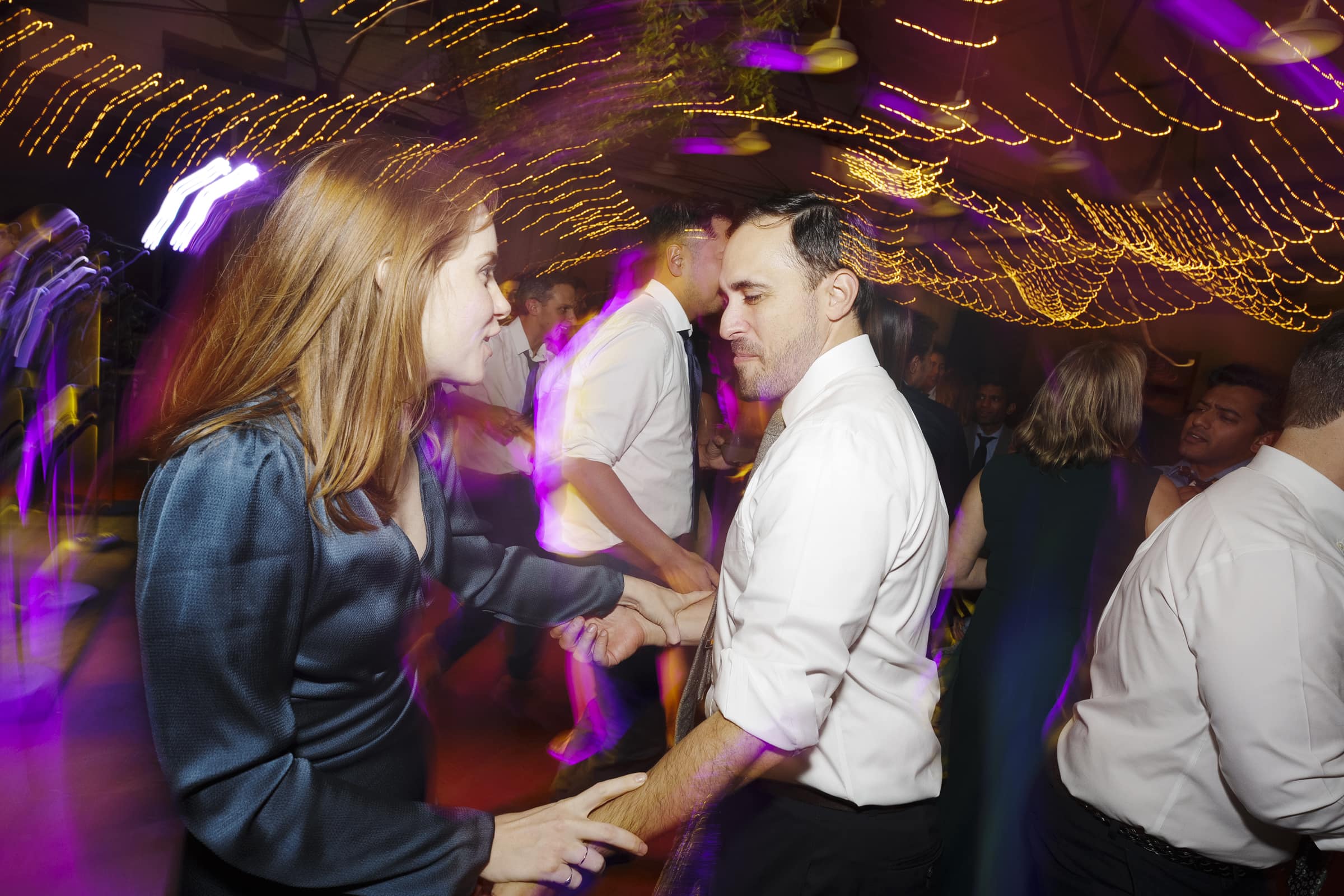 A group of people dance energetically under string lights at an indoor market hall wedding. A woman in a blue dress and a man in a white shirt are in focus, surrounded by purple and yellow light streaks, creating a lively atmosphere.
