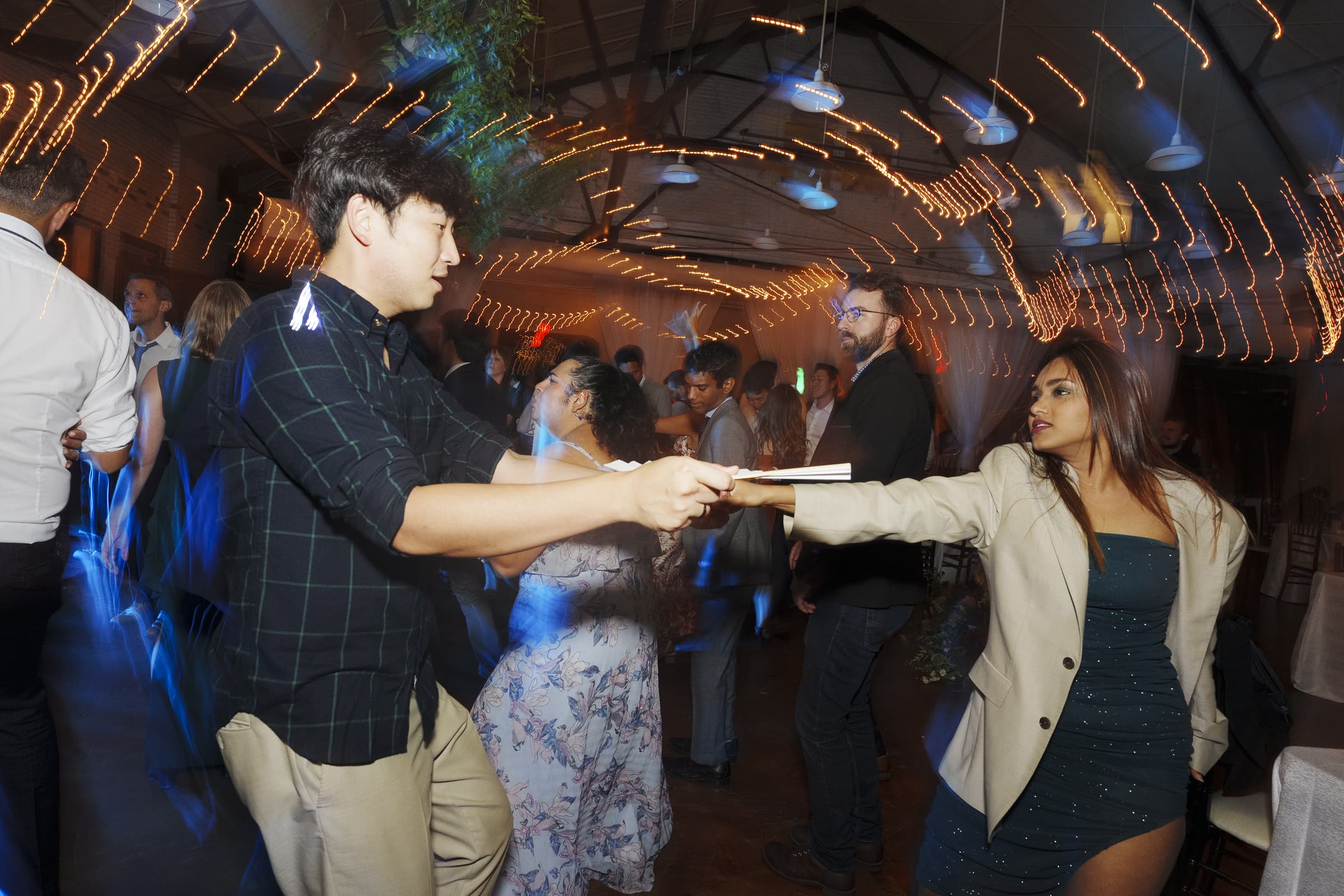 A man and woman dance energetically under string lights at a lively market hall wedding. The woman holds a baguette towards the man, adding a playful element. Other guests can be seen socializing in the softly lit background.