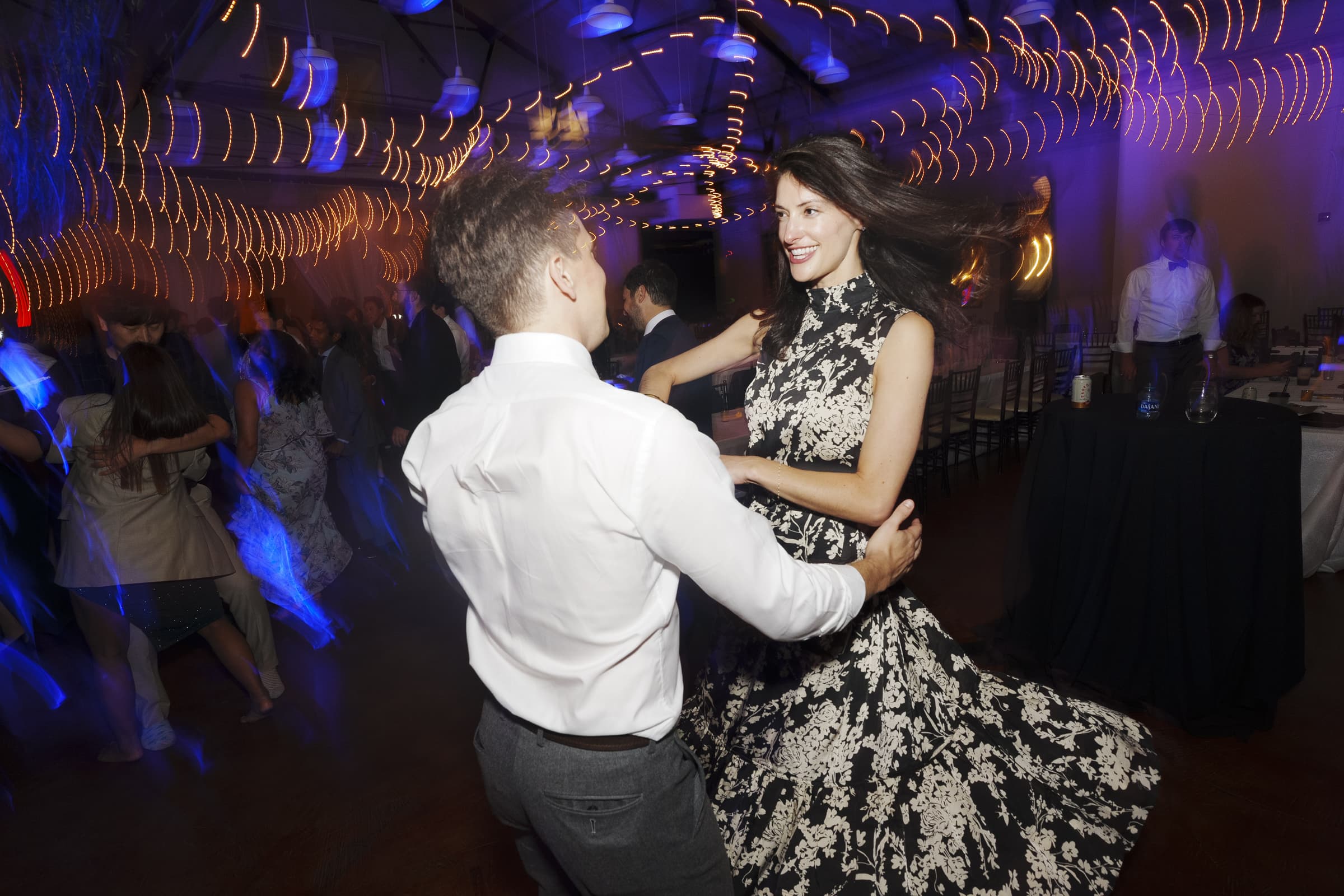 A couple dances energetically under string lights at a lively market hall wedding. The woman's dress swirls as she spins, while the colorful lighting enhances the festive atmosphere. Other guests are visible in the softly blurred background, adding to the vibrant scene.