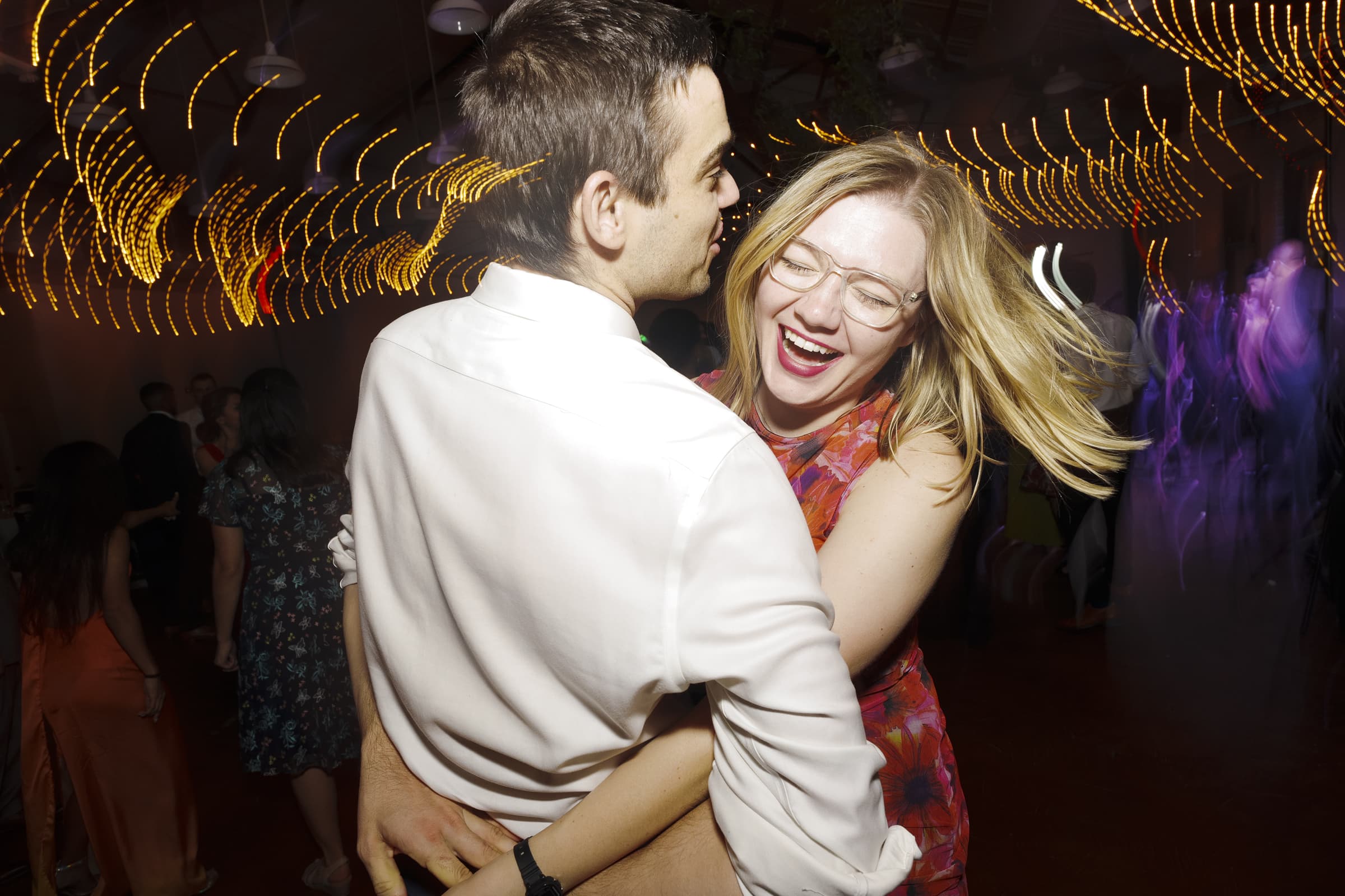 A couple dances joyfully under warm, swirling string lights at their market hall wedding. The woman, wearing glasses and a floral dress, laughs as her hair moves with the dance. The man in a white shirt embraces her, both reveling in the festive atmosphere.