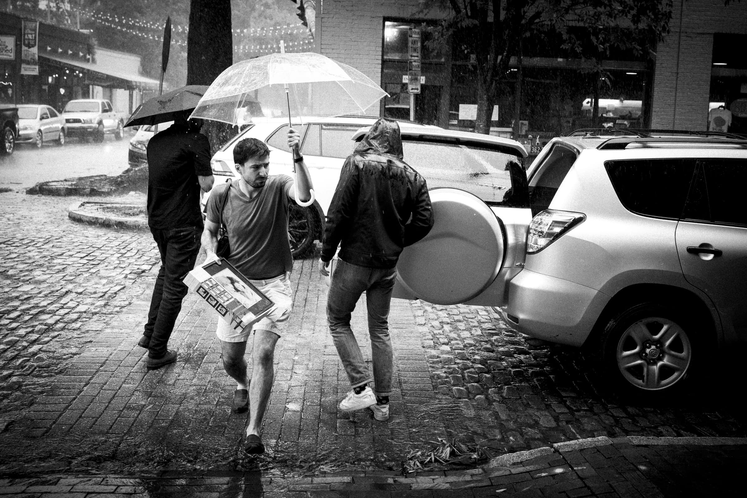 A black and white photo captures a trio caught in the rain on a cobblestone street. One person clutches an umbrella while carrying a box, perhaps for a market hall event; another grasps a large circular object near an open car trunk, as the third person strolls away with an umbrella.