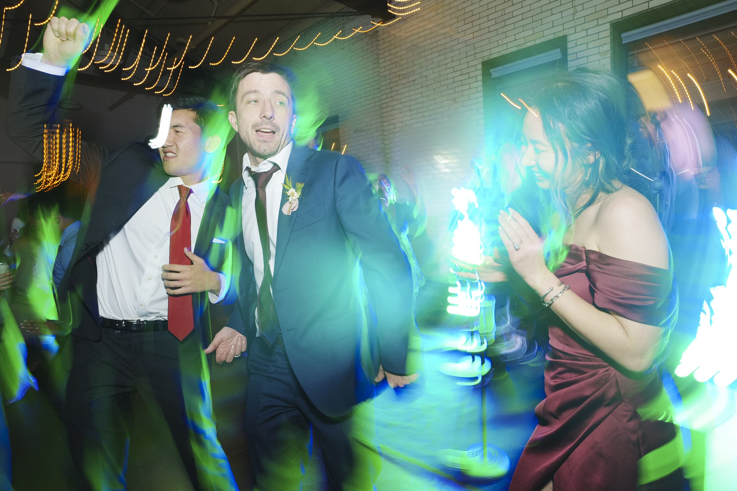 In a dimly lit market hall, three people in formal attire dance energetically, surrounded by colorful, blurred lights that create a lively wedding atmosphere. The person on the right twirls in a maroon dress, while the two on the left sport sharp suits.