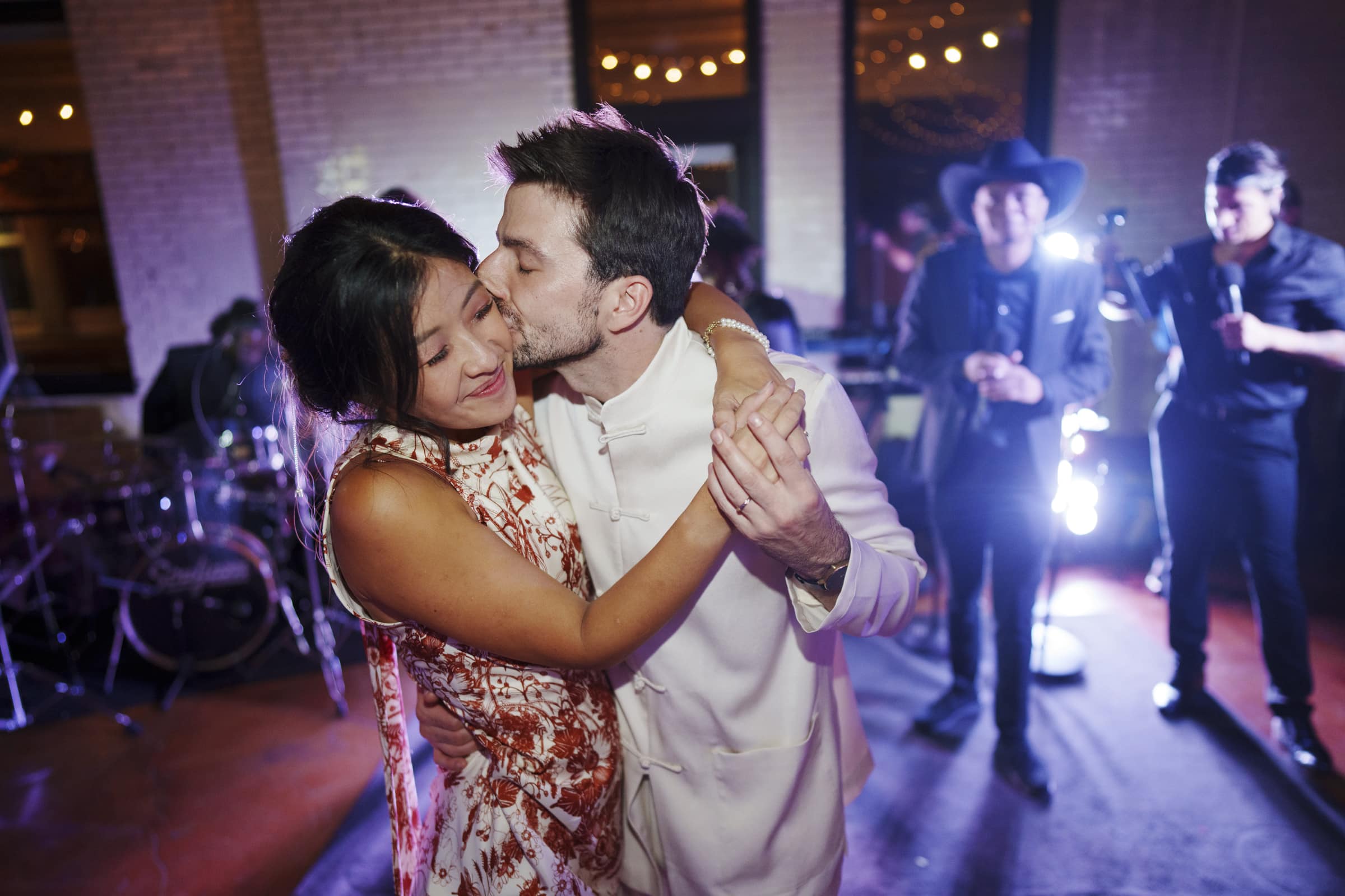A couple dances closely at a market hall wedding reception, with the man kissing the woman's cheek. They are surrounded by festive lighting and musicians performing in the background. The atmosphere is joyful and intimate.