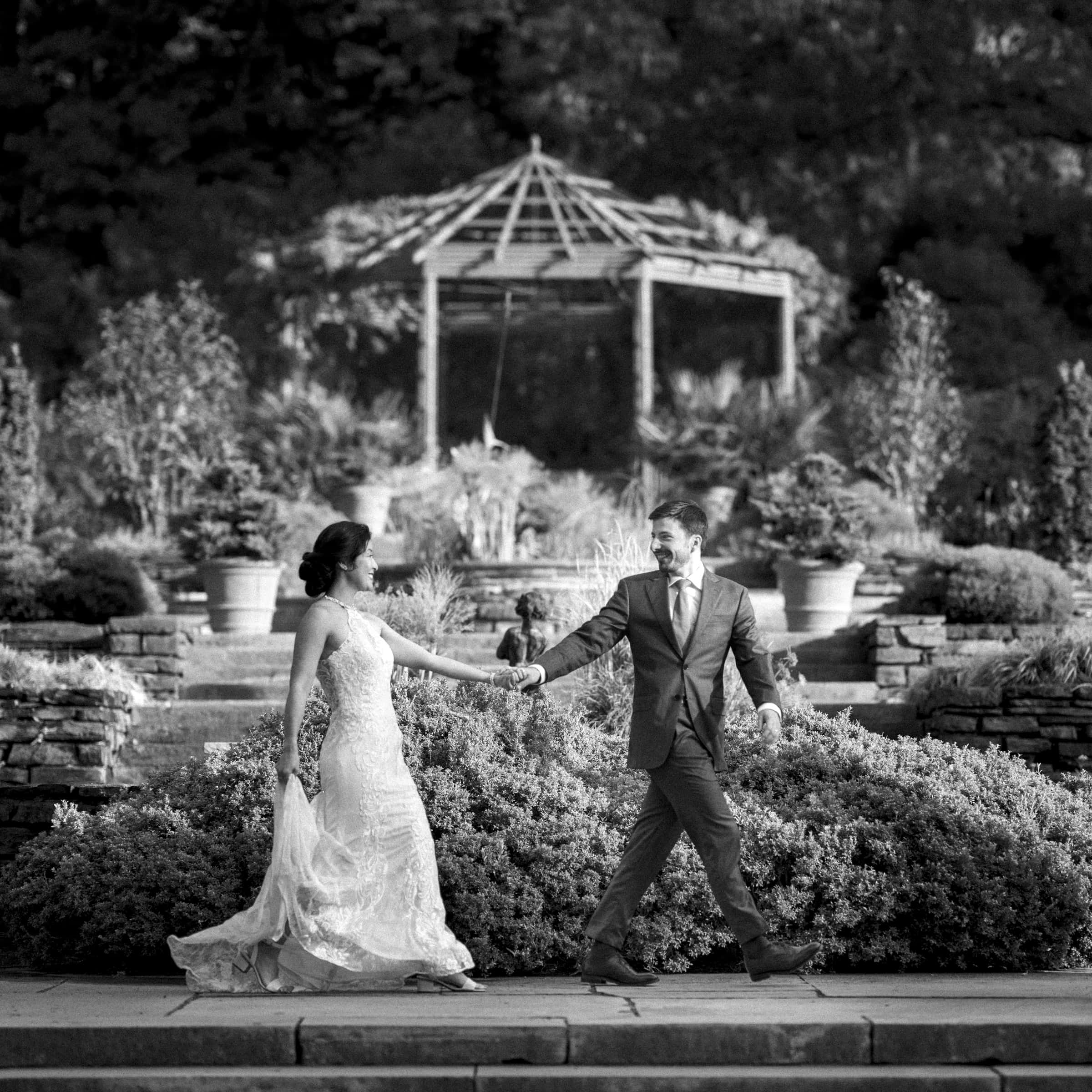 A bride and groom walk hand in hand outdoors, reminiscent of a market hall wedding. The bride wears a long, elegant gown, and the groom is in a suit. Surrounded by lush greenery with a gazebo in the background, the scene exudes timeless romance in black and white.