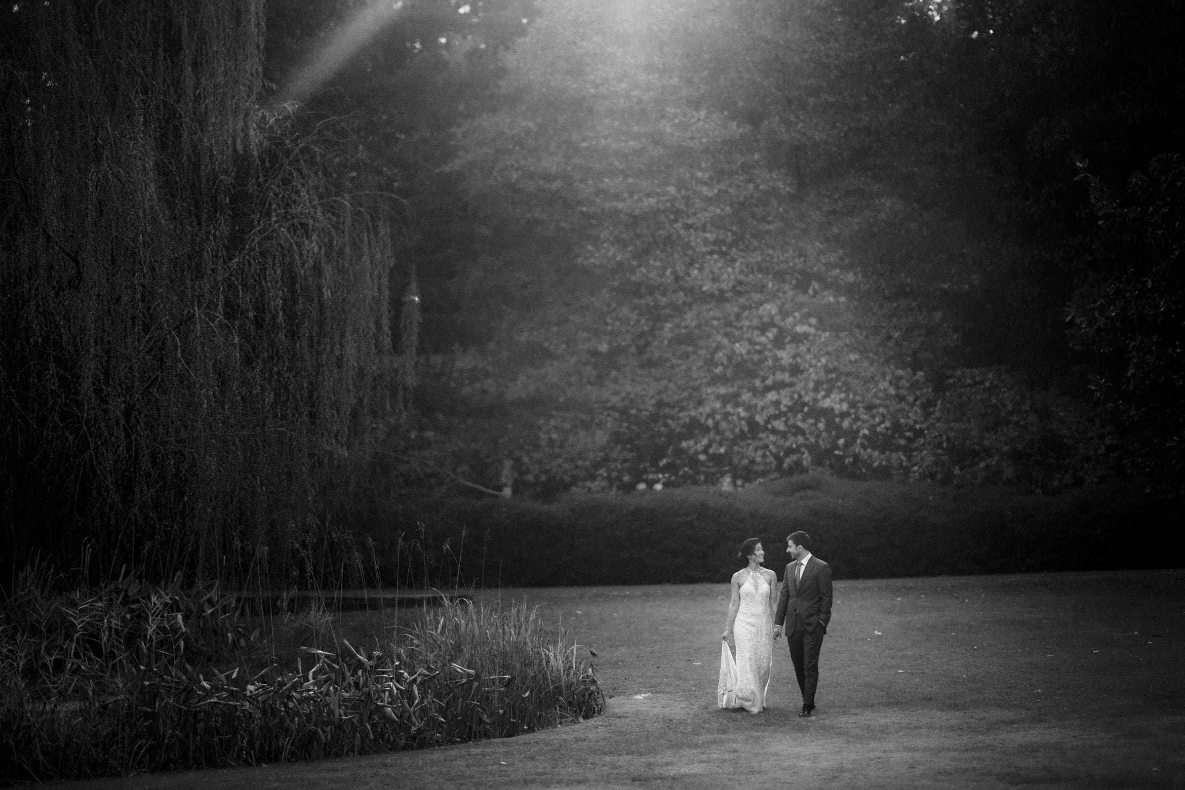 A bride and groom walk hand in hand on a grassy lawn at the market hall wedding venue, surrounded by trees. Sunlight filters through the foliage, casting a soft glow over the couple. The scene is serene and romantic, captured in black and white.