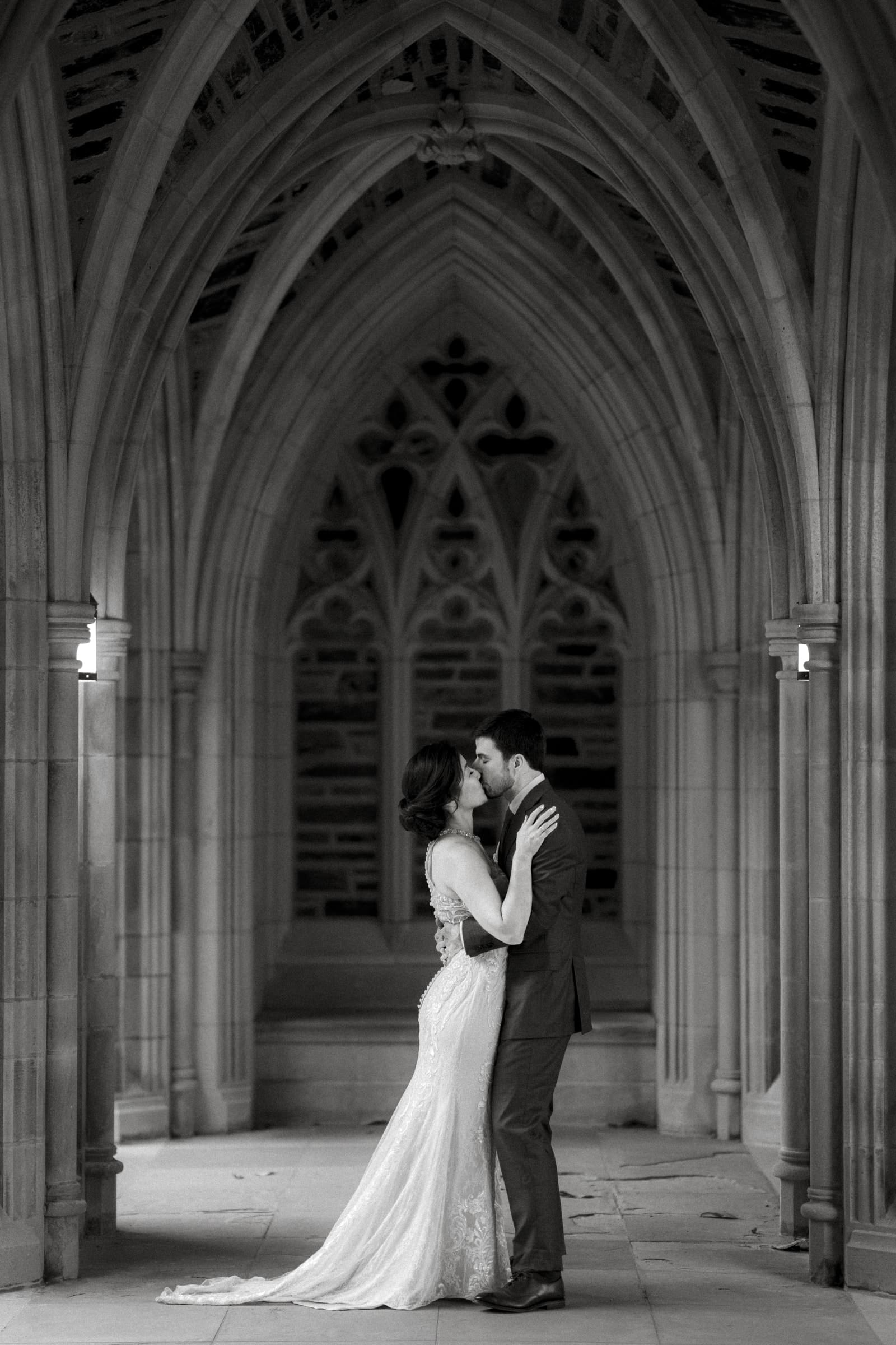 A couple stands close, embracing under the stone archways of a historic market hall in a black and white photo. The bride wears a long, elegant gown, and the groom is in a suit. The setting is ornate, with intricate architectural details that elevate their wedding's timeless charm.