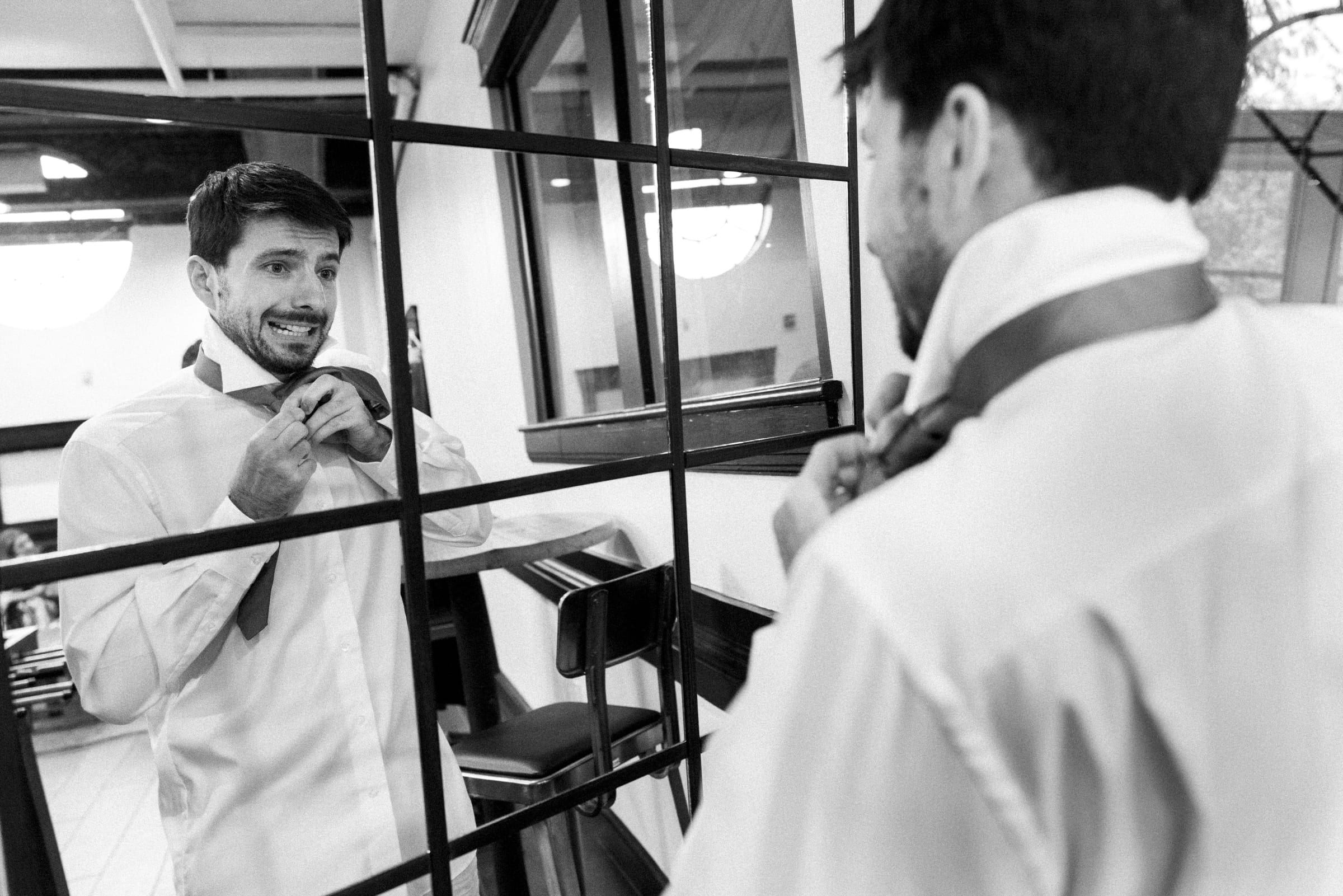 A man in a white shirt stands in front of a mirror, adjusting his tie with a focused expression, as if preparing for a market hall wedding. The scene is captured in black and white, situated in a well-lit room with a grid-like window reflection.