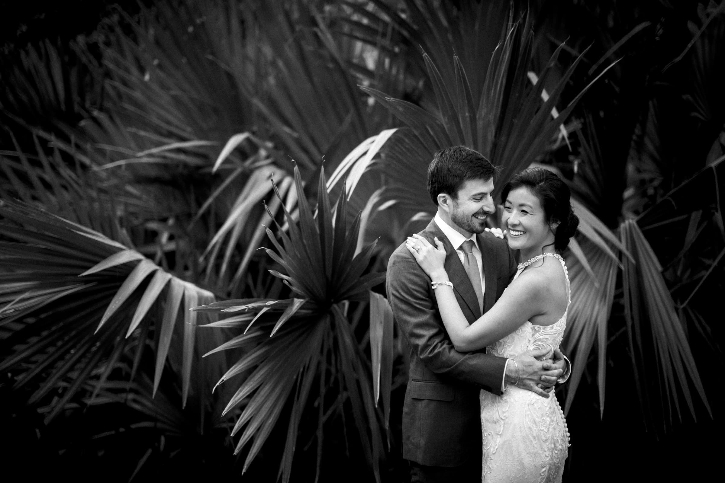 A black and white photo of a joyful couple embracing in front of large, leafy plants at a market hall wedding. The woman is wearing a detailed lace dress, and the man is in a suit. Both are smiling warmly, creating a romantic and elegant scene.