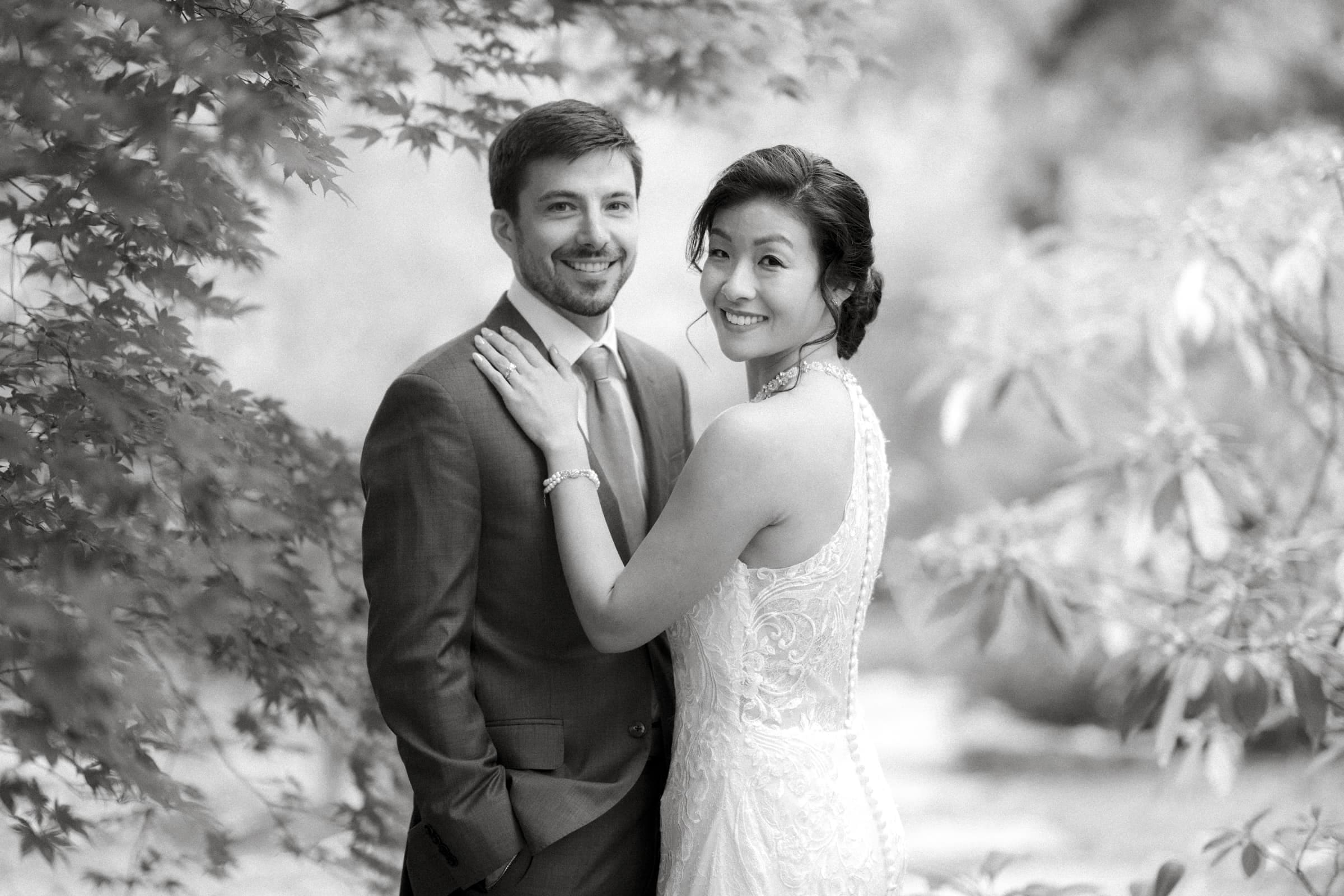 A black and white photo of a smiling couple in a garden setting, possibly after their market hall wedding. The woman, in a detailed lace dress, rests her hand on the man's shoulder. He wears a suit and tie. Both look at the camera, surrounded by leafy branches.