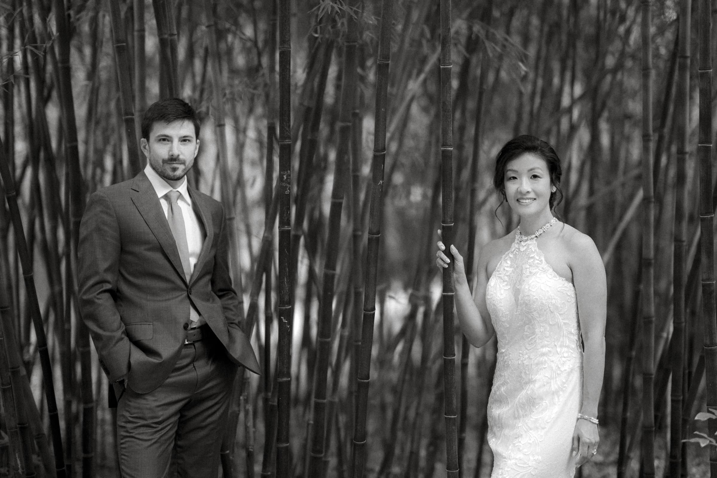 A man in a suit and a woman in a sleeveless white dress stand among tall bamboo, exuding the charm of an intimate market hall wedding. The man has his hands in his pockets, while the woman gently holds a bamboo stalk. Both face the camera with soft smiles, surrounded by lush greenery.