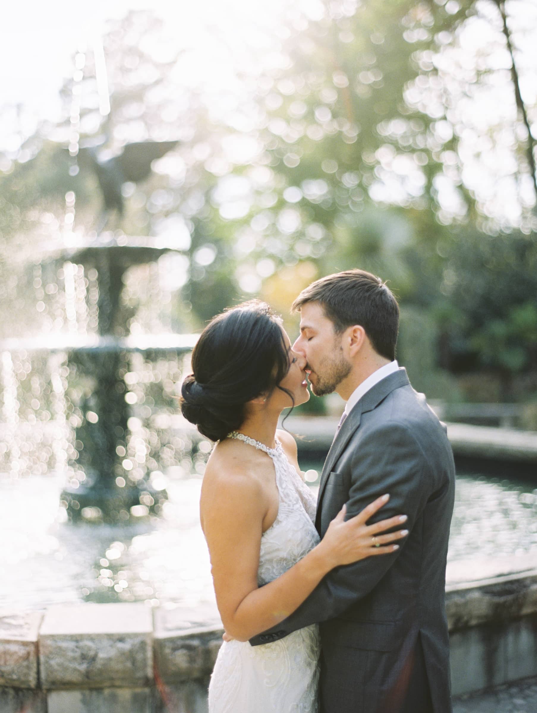 A couple in wedding attire shares a kiss in front of a sunlit fountain at their market hall wedding. The bride shines in a white lace gown, while the groom looks dapper in his gray suit. The backdrop is adorned with trees and a soft, glowing light, creating an enchanting scene.