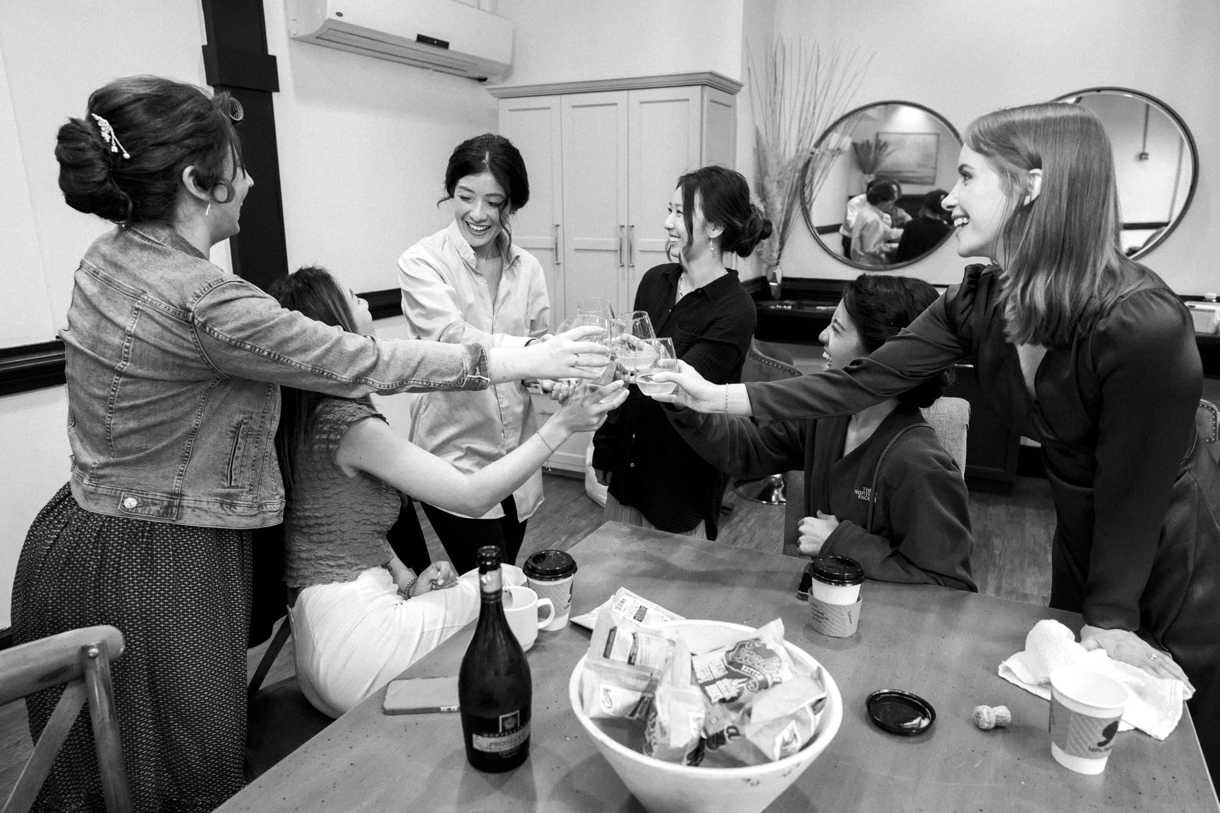 A group of six people cheerfully clinks glasses at a cozy market hall, surrounded by snacks and drinks. The indoor setting features round mirrors and a cabinet in the background. Everyone appears to be enjoying themselves, celebrating what seems to be a lively wedding gathering.