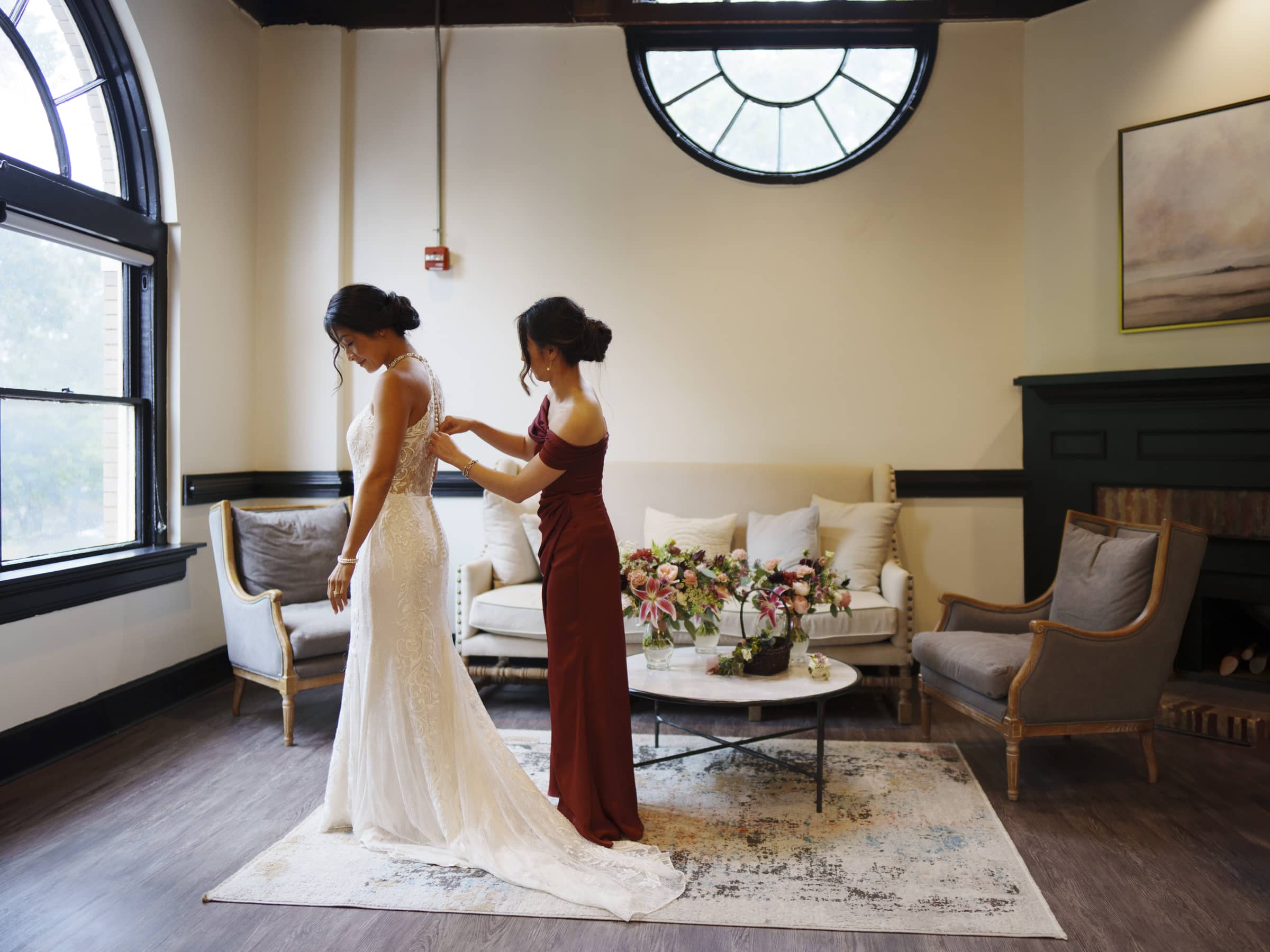 In a beautifully adorned room reminiscent of a market hall wedding, a bride in a white gown is assisted by another woman in a burgundy dress. Elegant furniture surrounds them, while a bouquet of flowers rests gracefully on the coffee table beneath large round windows.