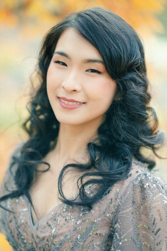 A woman with long, wavy black hair smiles softly, wearing a glittery, sheer top during her Duke University engagement session. The background is a soft blur of autumn colors, suggesting an outdoor setting.