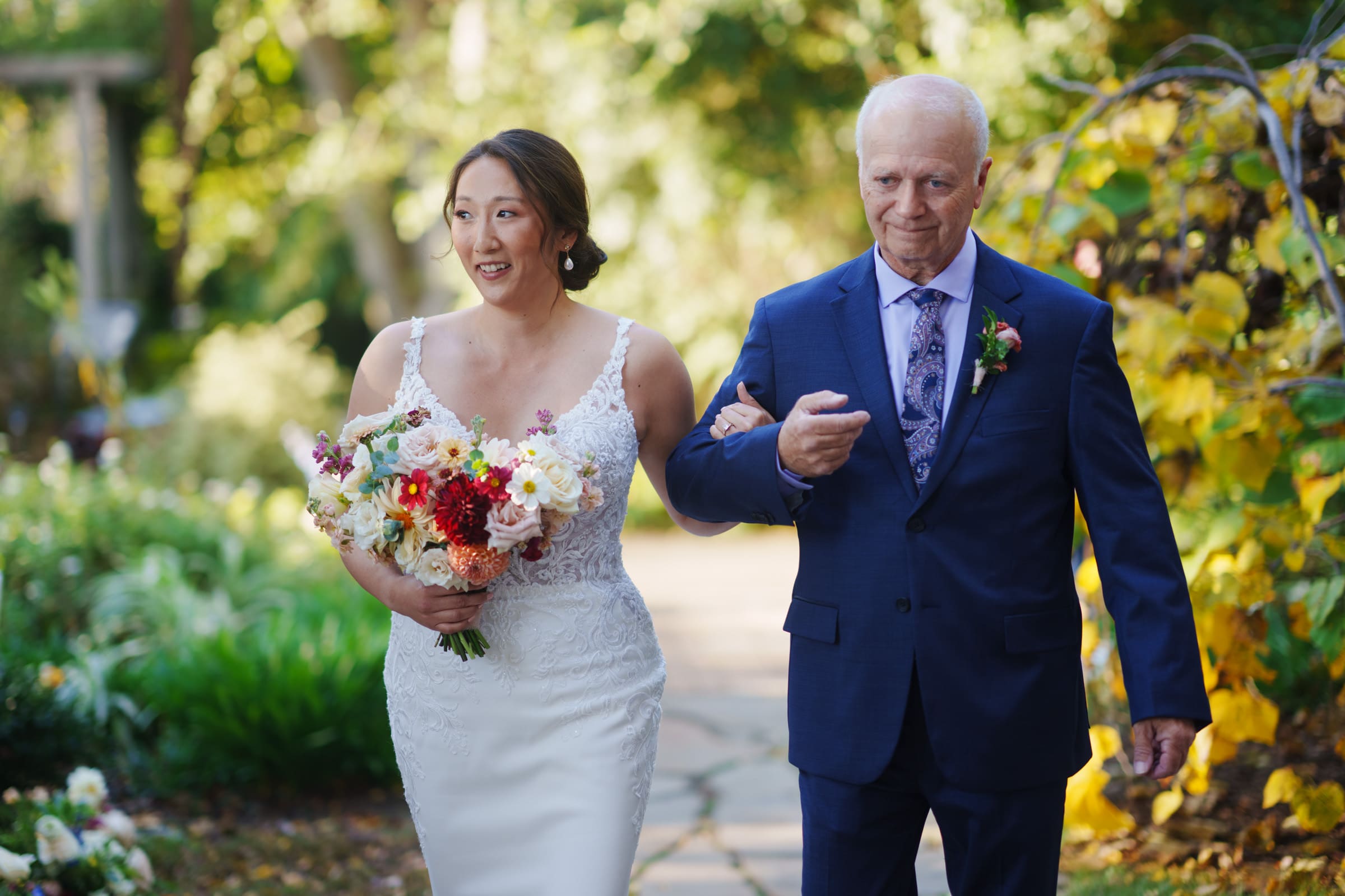 A bride in a white lace gown holds a vibrant bouquet as she walks arm-in-arm with an elderly man in a blue suit. They gracefully navigate the garden path at JC Raulston Arboretum, surrounded by greenery and yellow foliage, capturing the essence of Holly & Sut's wedding day.