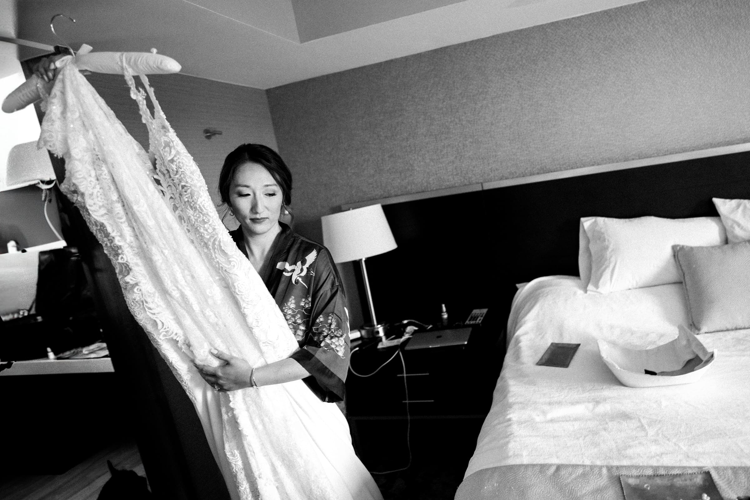 A woman in a robe admires a lace wedding dress on a hanger in her modern, neutral-toned bedroom. The setting is serene and elegant, reminiscent of the style seen at Holly & Sut's JC Raulston Arboretum wedding. Beside her, a lamp softly glows on the nightstand.