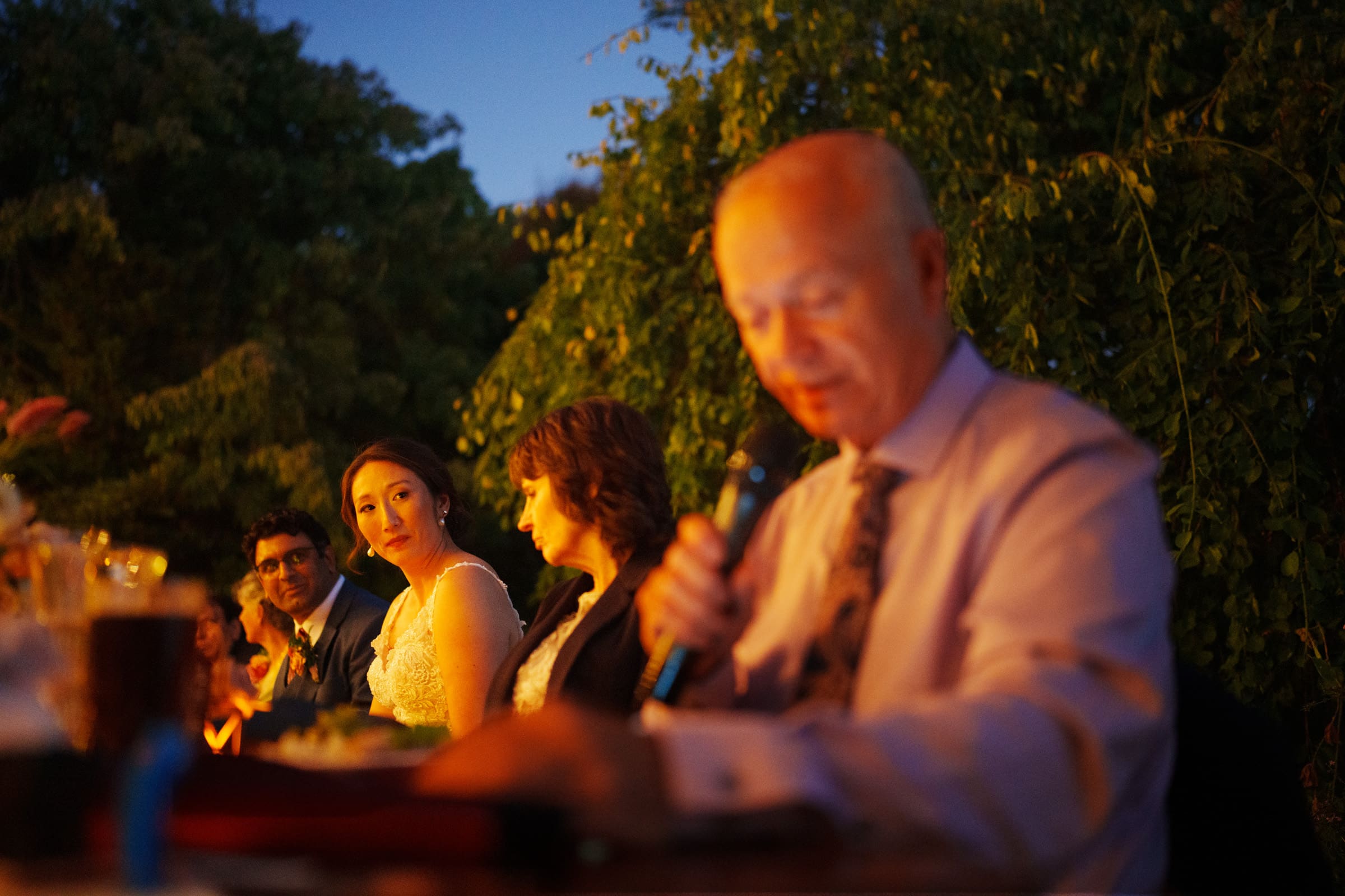 At a charming JC Raulston Arboretum wedding, Holly gazes lovingly at Sut under the warm glow of lights. An older man in the foreground holds a microphone, likely delivering a heartfelt speech, as tables of guests enjoy the serene outdoor setting framed by lush trees.