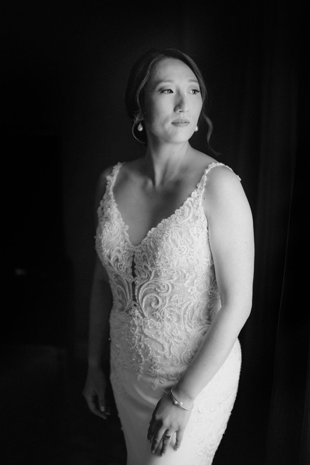 A woman in a lace wedding dress stands near a window at the JC Raulston Arboretum, gazing out thoughtfully. The black and white image highlights the intricate details of her gown and her serene expression—perfectly capturing the elegance of Holly & Sut's special day.