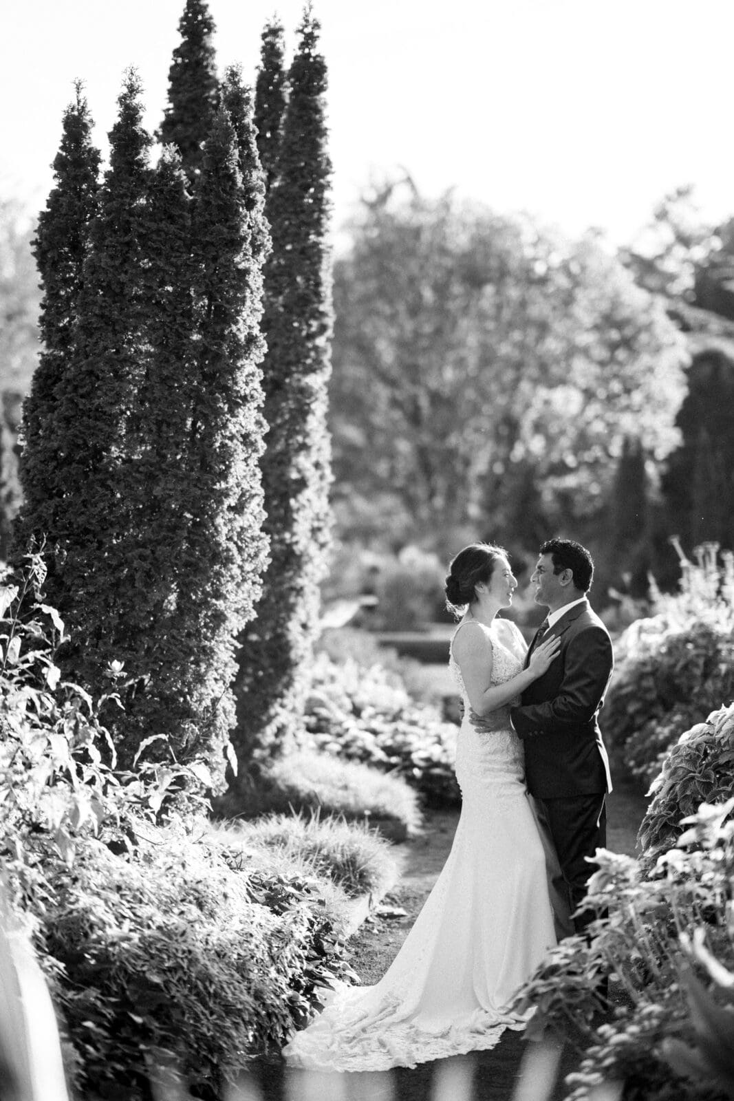 A black and white photo captures Holly and Sut in formal attire embracing on a garden path at JC Raulston Arboretum. Tall trees and lush foliage surround them, with sunlight softly illuminating the scene. The couple gazes into each other's eyes, creating a romantic atmosphere.