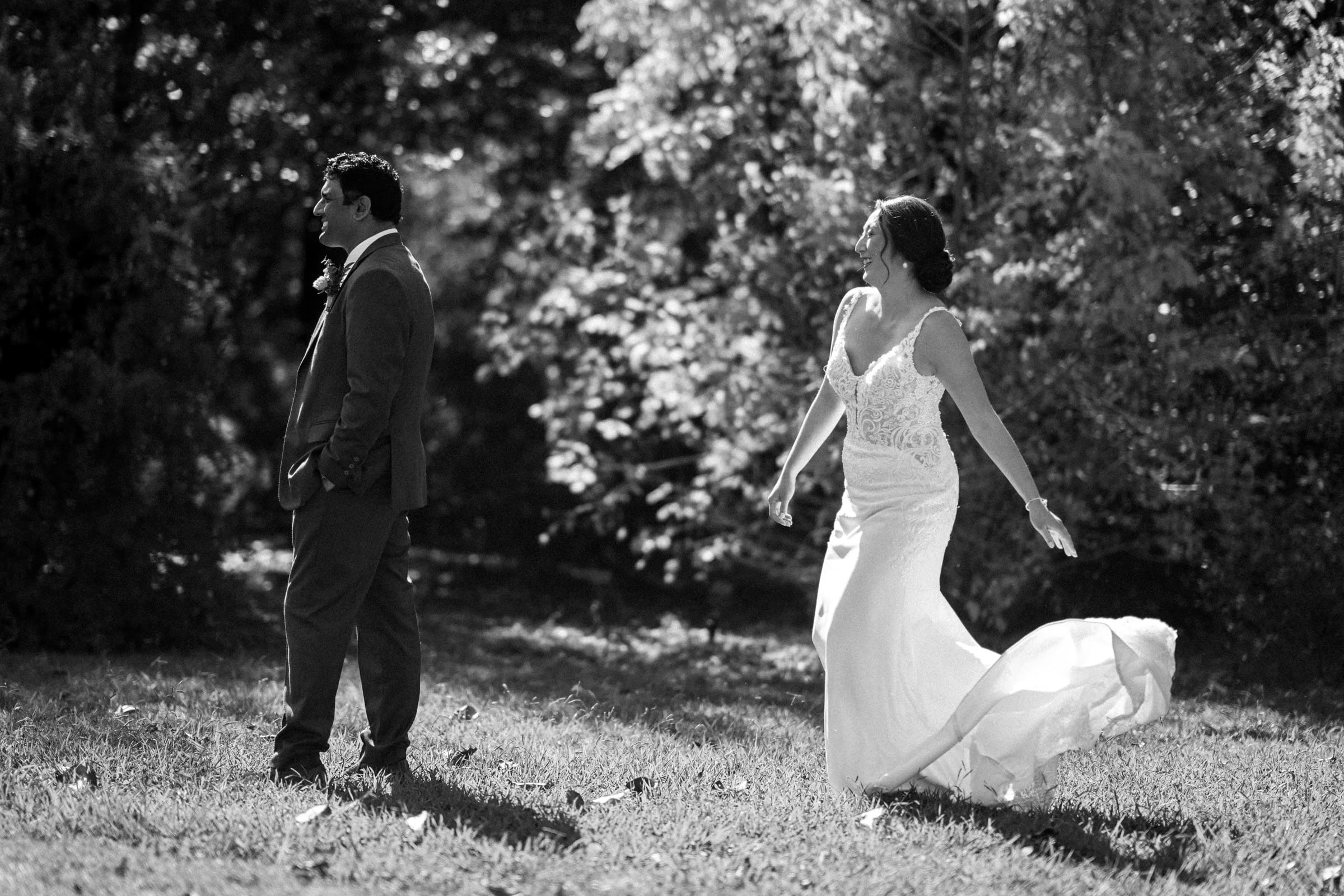 A black-and-white photo captures Holly in a flowing gown walking towards Sut at their JC Raulston Arboretum wedding. The bride's dress sways gracefully as she approaches the groom, standing on a grassy area with trees in the background.