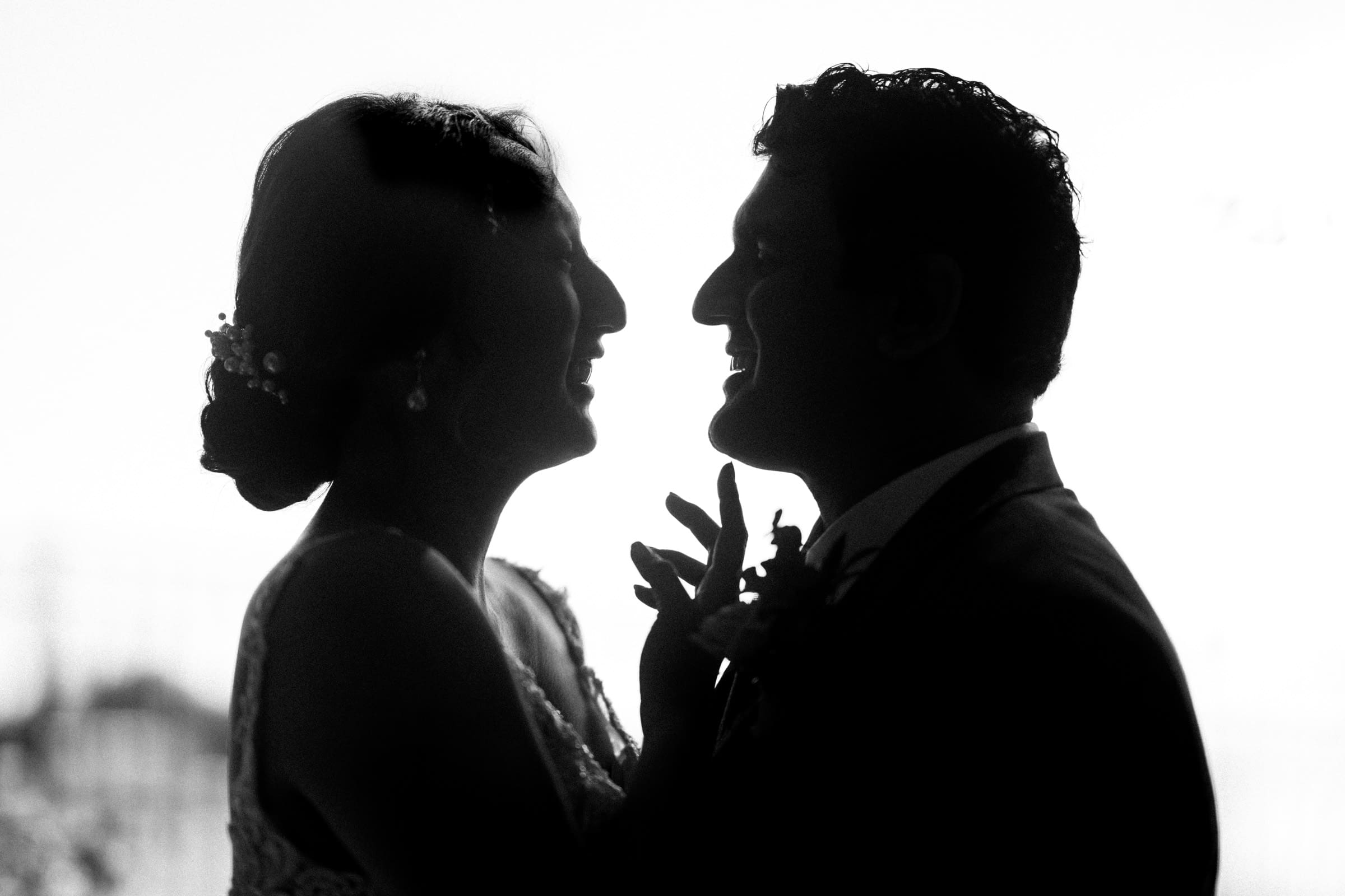 A black and white silhouette of Holly and Sut, a joyful couple at their JC Raulston Arboretum wedding, smiling as she, with her hair in an updo adorned with flowers, touches his chest. Both are elegantly dressed in formal attire against a softly out-of-focus background.