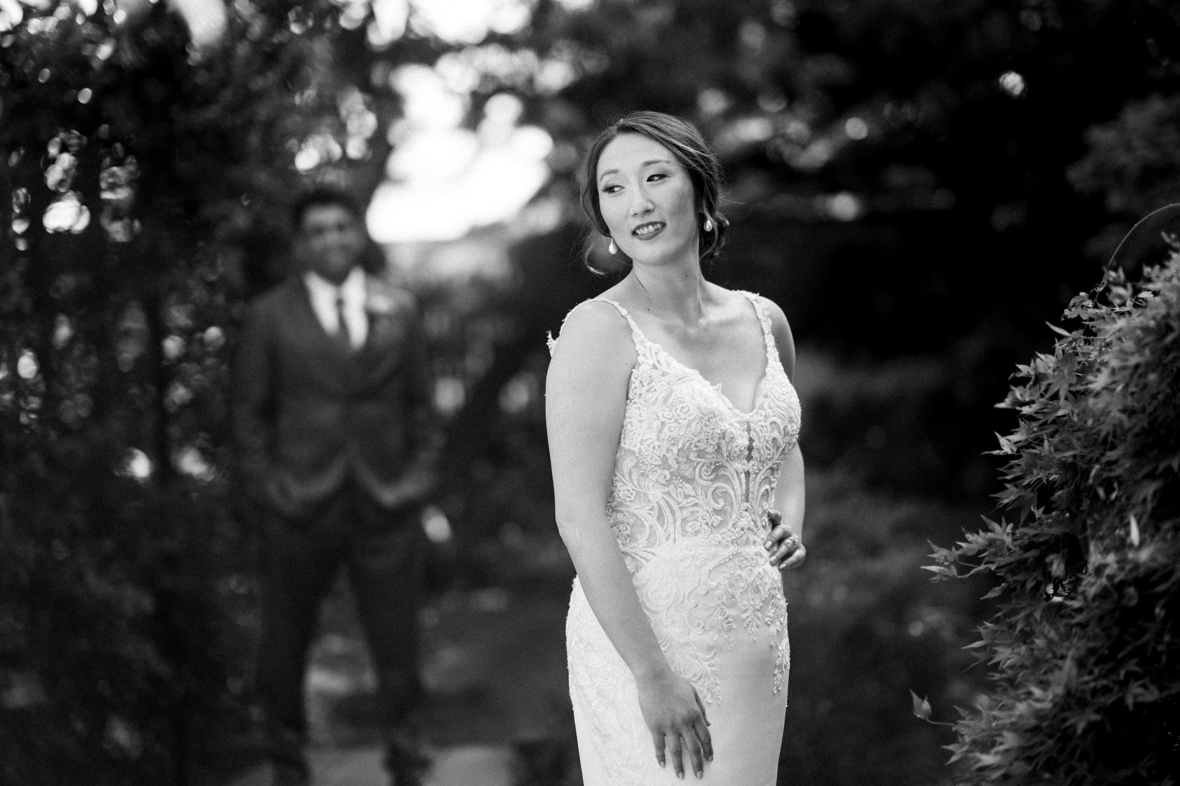 A black and white photo captures a bride in a lace wedding dress at the JC Raulston Arboretum, looking to the side amid lush greenery. In the background, a groom in a suit—perhaps Holly’s beloved Sut—is slightly blurred, tenderly gazing at her.