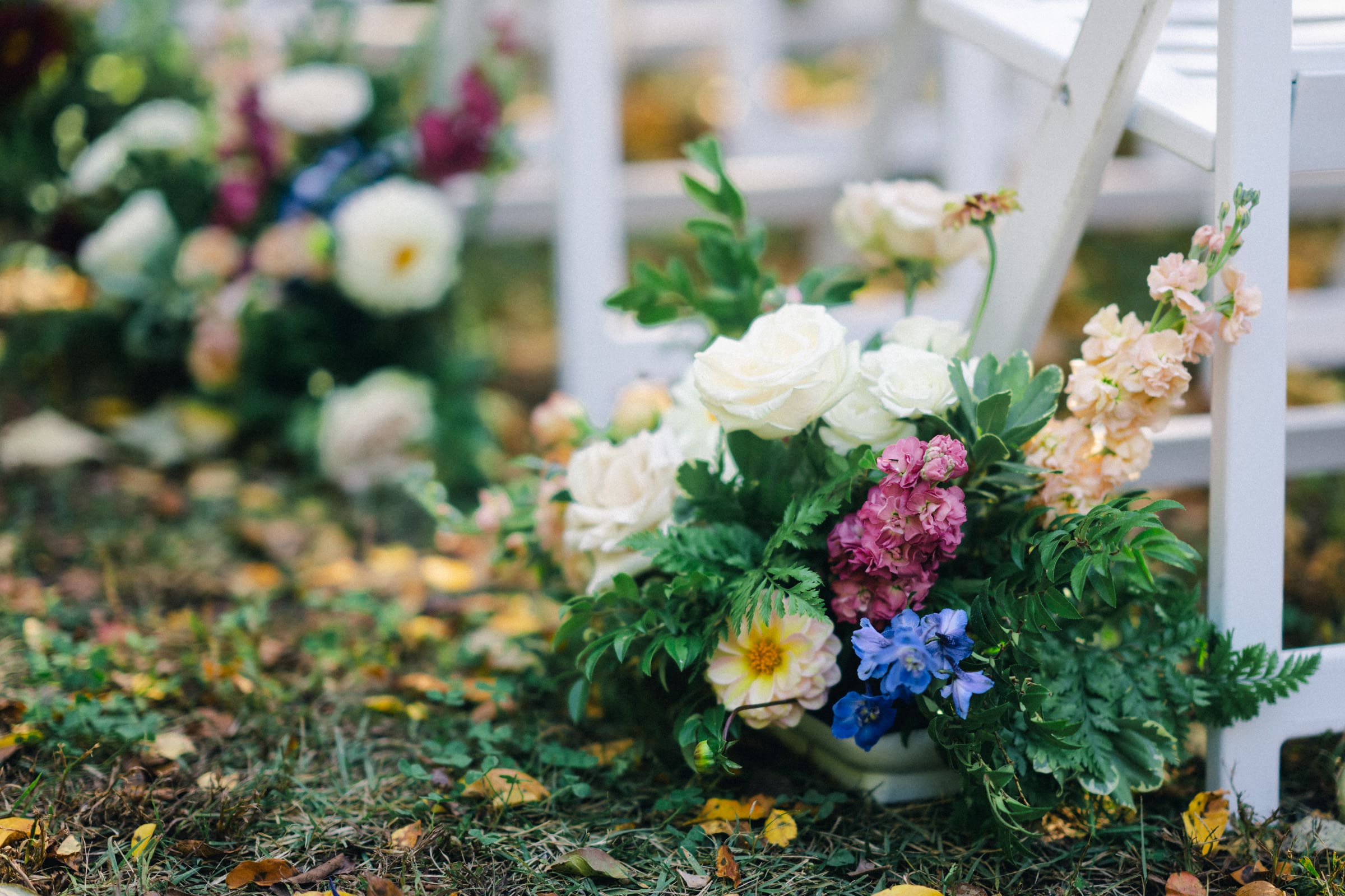 A decorative floral arrangement featuring white roses, pink and blue flowers, and lush greenery graces a JC Raulston Arboretum wedding. Beside a white wooden chair, the display rests on grass sprinkled with scattered autumn leaves—a perfect setting for Holly & Sut's special day.