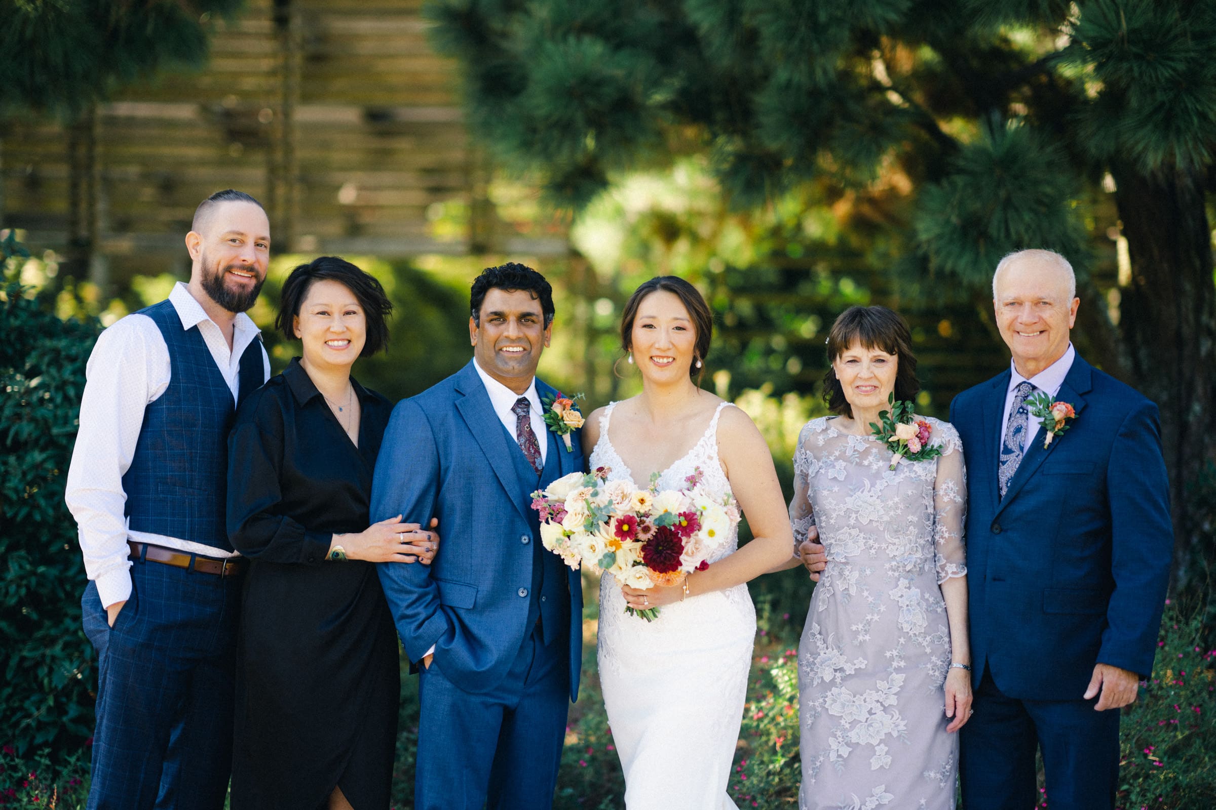 A group photo of six people standing outdoors in formal attire at the JC Raulston Arboretum wedding of Holly & Sut. The bride holds a bouquet, and others are wearing suits and dresses. They are smiling, with greenery and a wooden fence in the background.