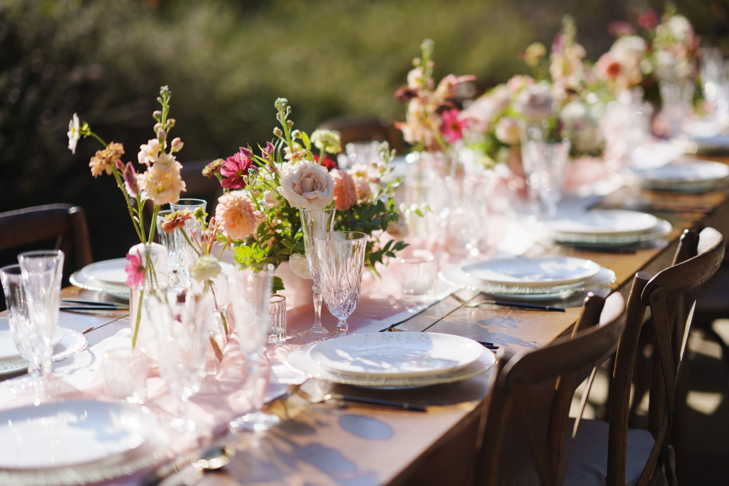 A beautifully set outdoor table at the JC Raulston Arboretum wedding of Holly & Sut features elegant plates, crystal glasses, and floral centerpieces with pink and white flowers. The warm sunlight adds a natural glow, creating an inviting and festive atmosphere.