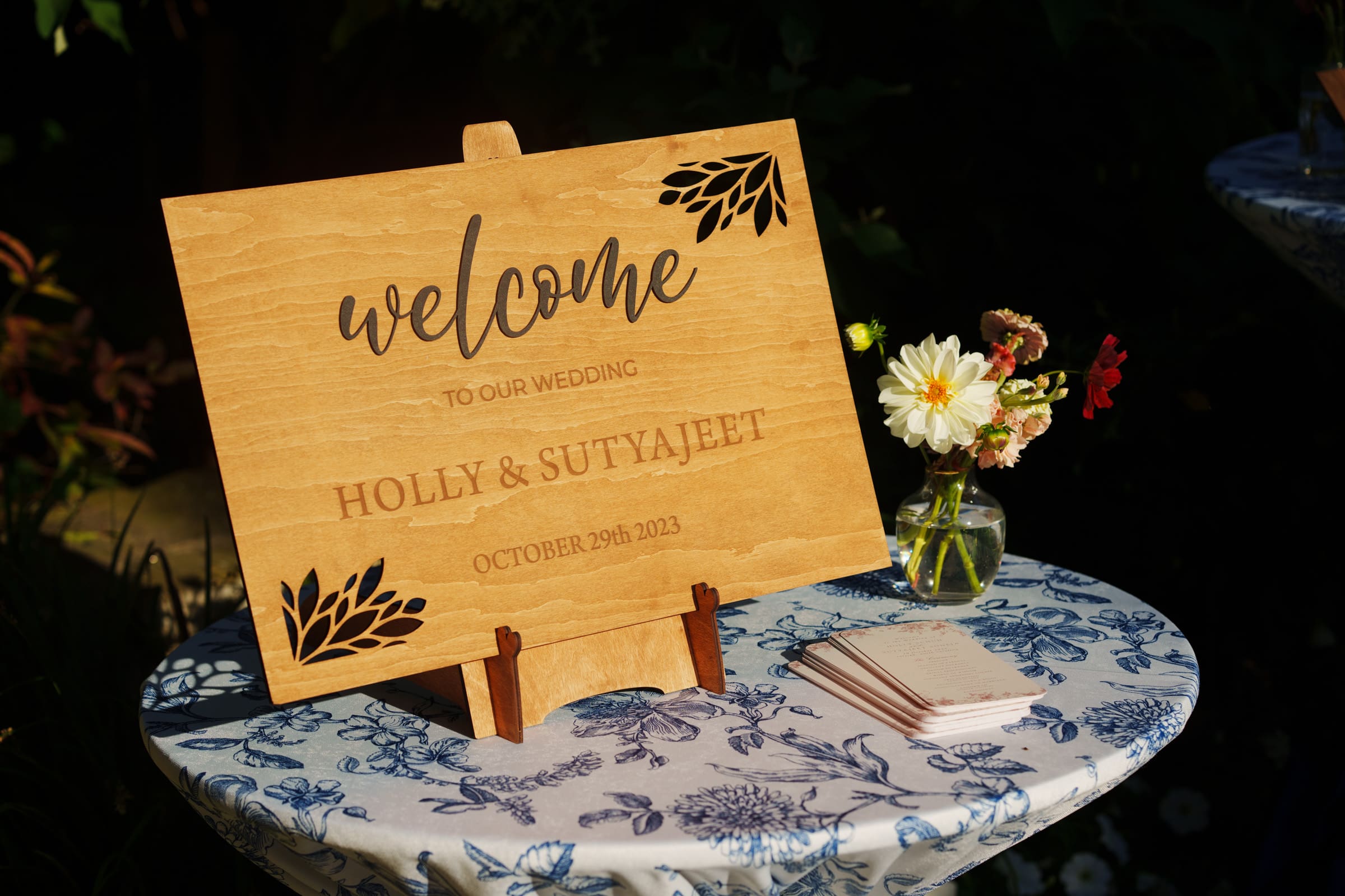 A wooden sign on a table reads, "Welcome to our wedding, Holly & Sutyajeet, October 29th, 2023," adorned with exquisite floral motifs. Celebrating at the JC Raulston Arboretum Wedding, the floral cloth-draped table is embellished with blooms and neatly stacked napkins.
