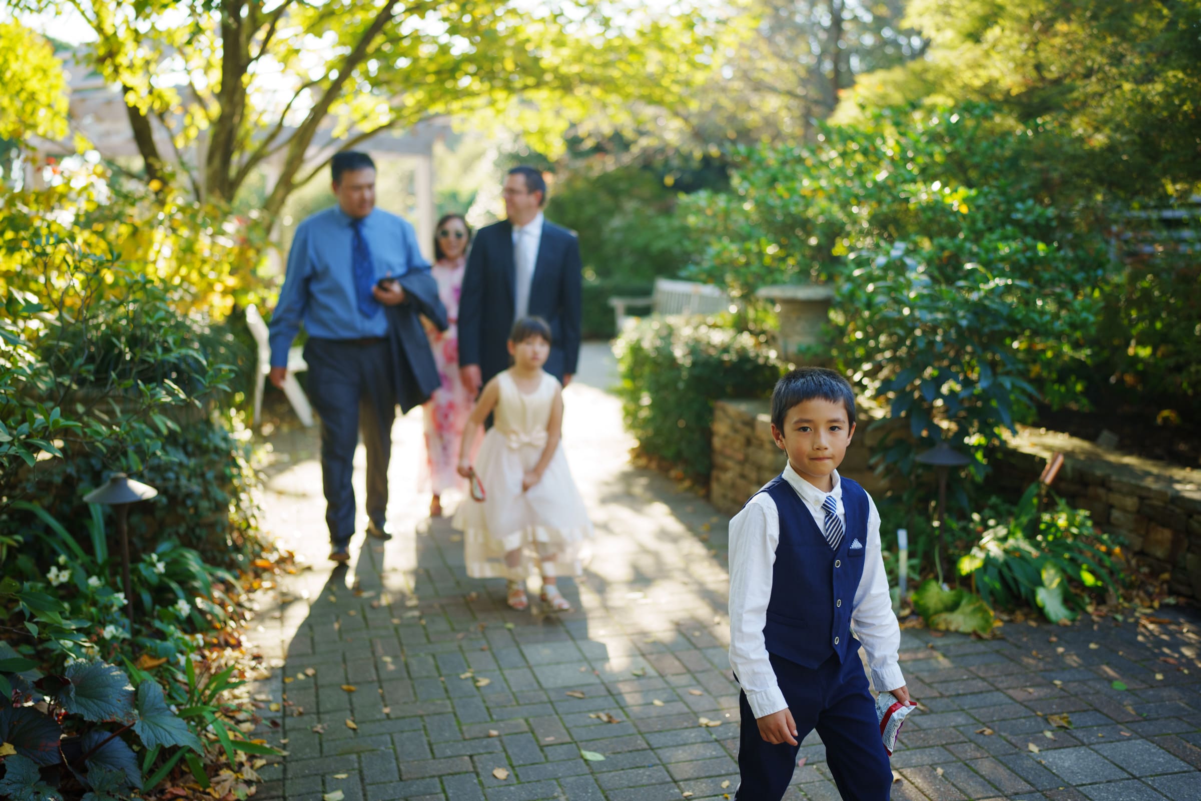 A young boy in a suit walks along the garden path at the JC Raulston Arboretum wedding of Holly & Sut, holding a card. Behind him, two men in suits and two girls in dresses follow, surrounded by lush greenery and soft sunlight filtering through the trees.
