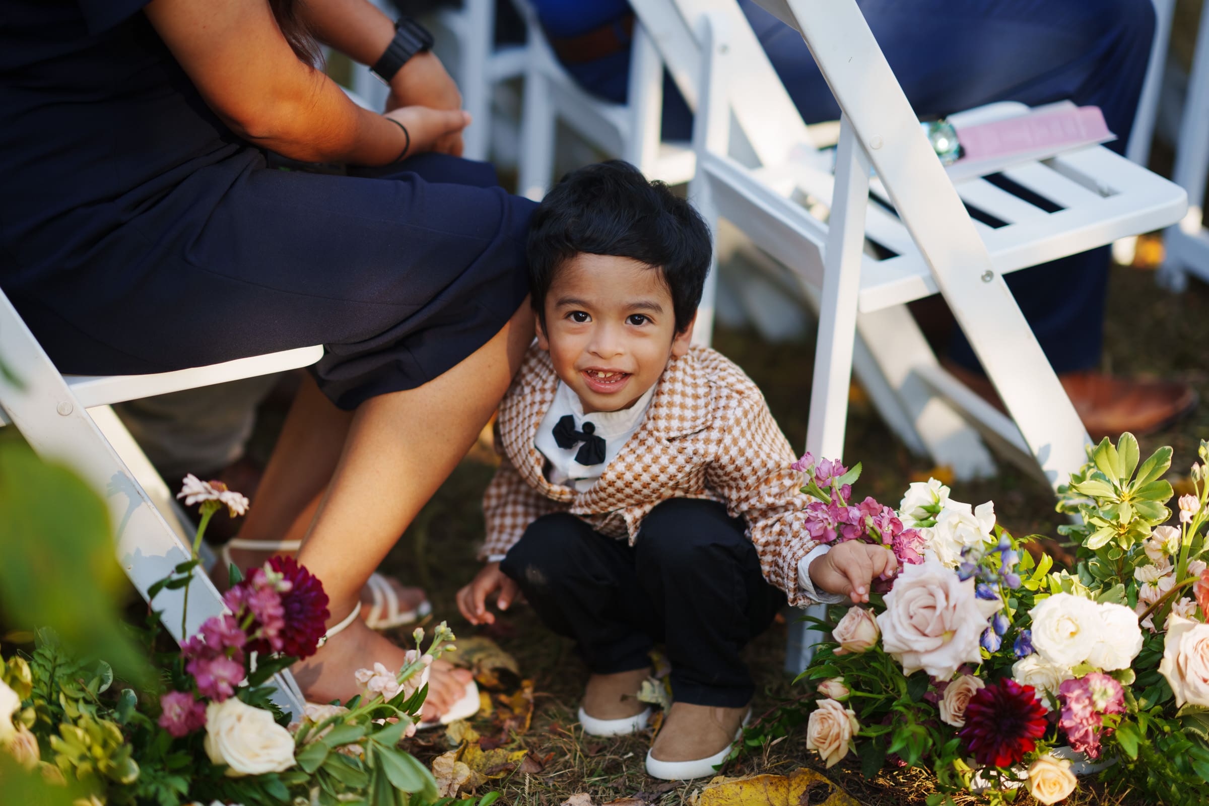 At the JC Raulston Arboretum wedding, a young child in a stylish outfit crouches beside a woman's legs. The child smiles at the camera, surrounded by vibrant flowers and white chairs, with sunlight creating a warm atmosphere. It’s a joyous celebration of Holly & Sut's special day.