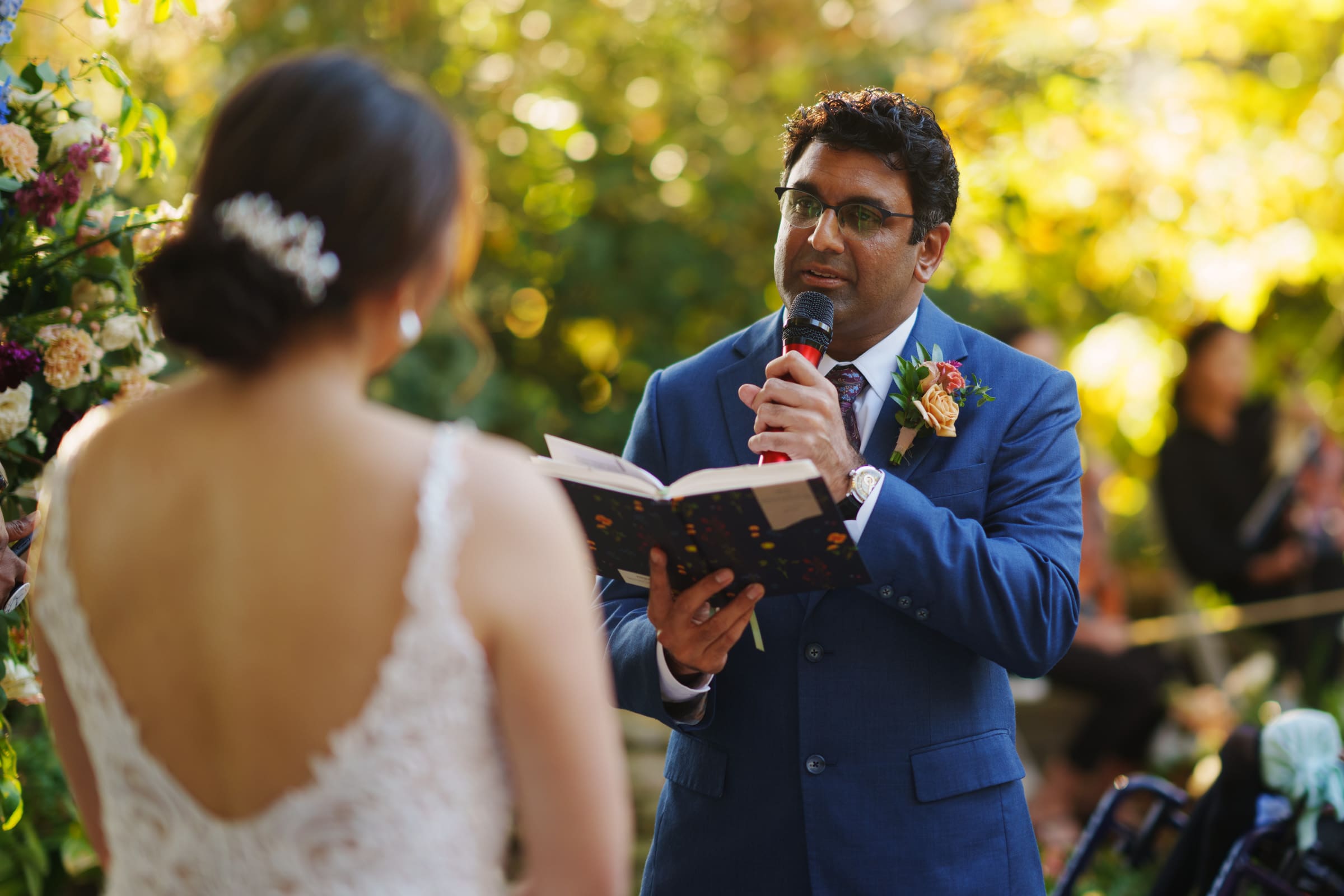 A man in a blue suit speaks into a microphone, holding a book, facing a woman in a white lace dress with a floral hairpin. They are at Holly and Sut's JC Raulston Arboretum wedding, surrounded by lush greenery and blooming flowers under the sunlit sky.
