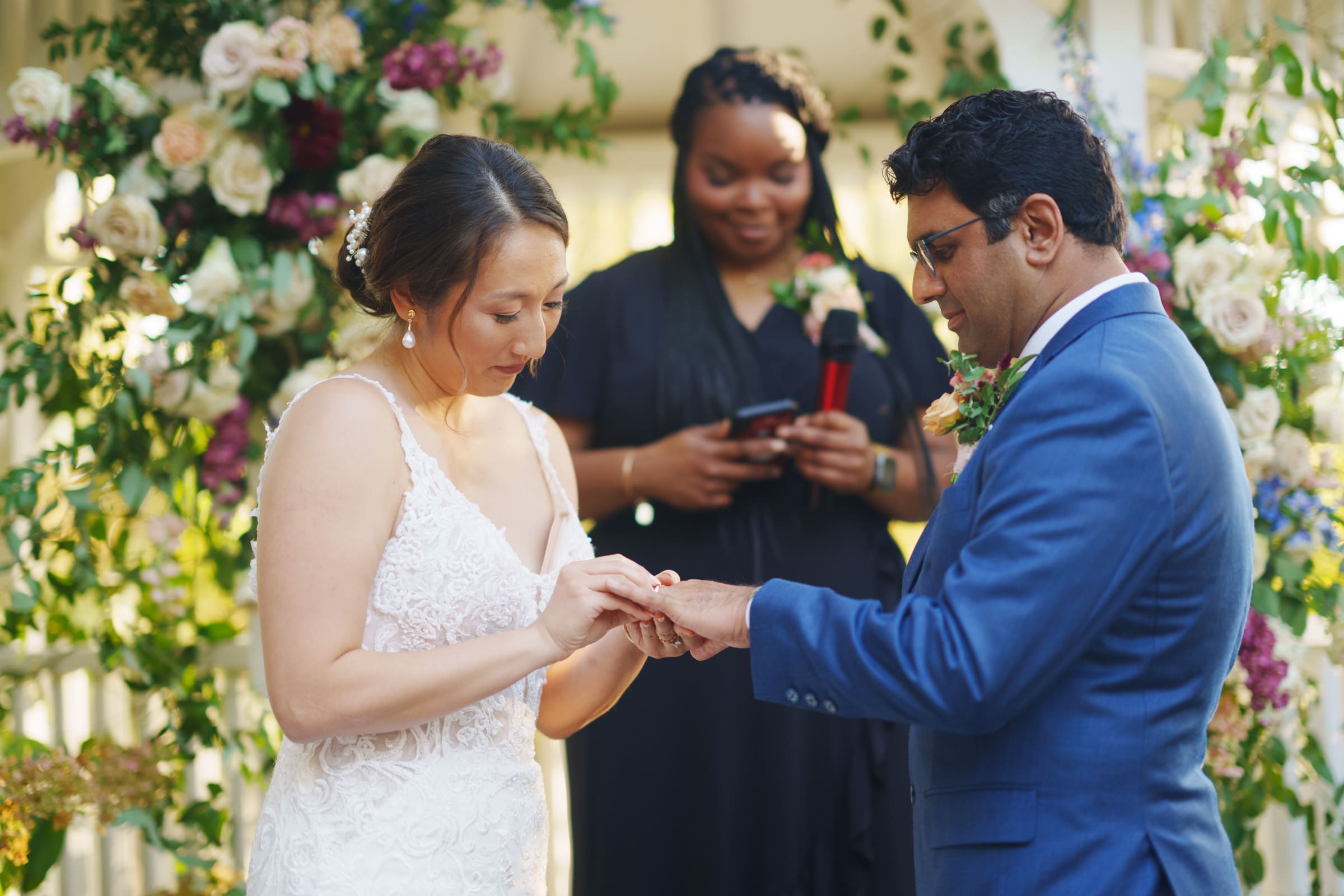 At the JC Raulston Arboretum wedding, Holly delicately places a ring on Sut's finger. She radiates elegance in her white dress while he looks dashing in a blue suit. The officiant, dressed in black, stands before a stunning floral backdrop, capturing the essence of their special day.