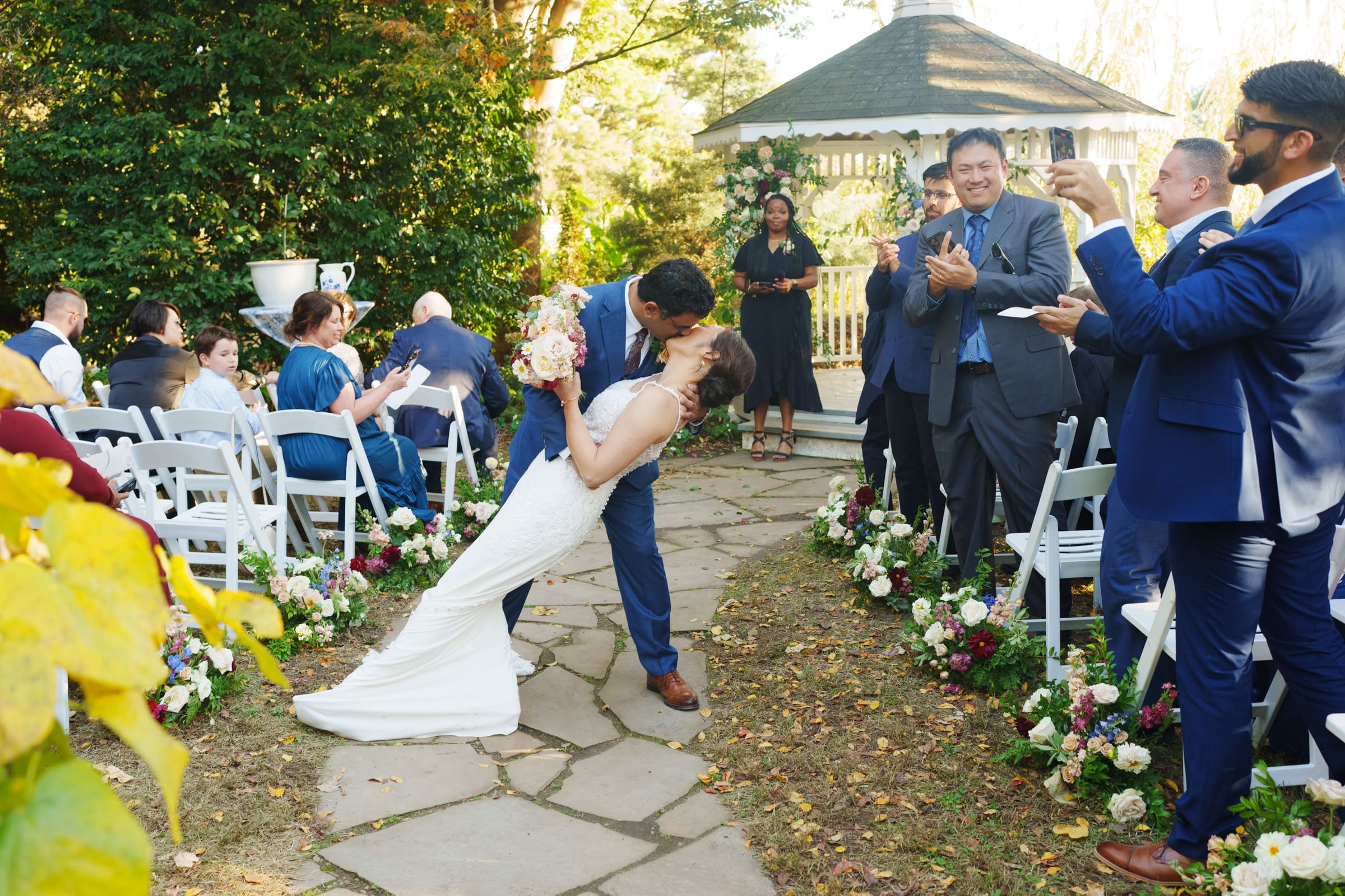 A newlywed couple, Holly and Sut, share a romantic kiss on a stone path at their stunning JC Raulston Arboretum wedding. Guests seated in white chairs watch and applaud. A gazebo and lush greenery serve as the backdrop, with flower arrangements lining the aisle.