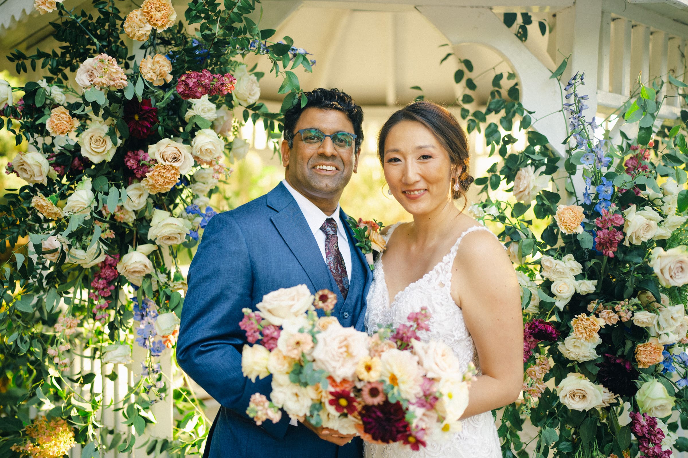 A bride and groom, Holly and Sut, stand smiling in front of a floral arch at the JC Raulston Arboretum wedding. The bride holds a bouquet of assorted flowers and wears a white lace gown, while the groom is in a blue suit with a patterned tie. The setting is an outdoor garden with lush greenery.