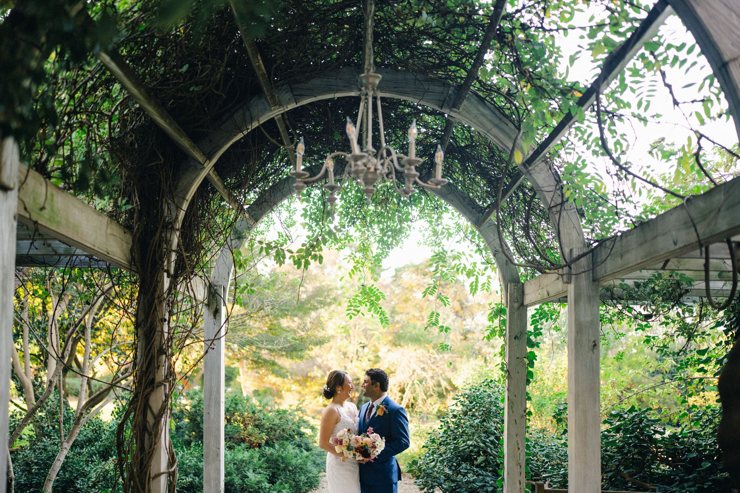 Holly and Sut stand under a vine-covered archway at the JC Raulston Arboretum, lost in each other's eyes. The bride clutches a bouquet beneath a chandelier, enhancing the lush, green garden setting of their wedding venue.