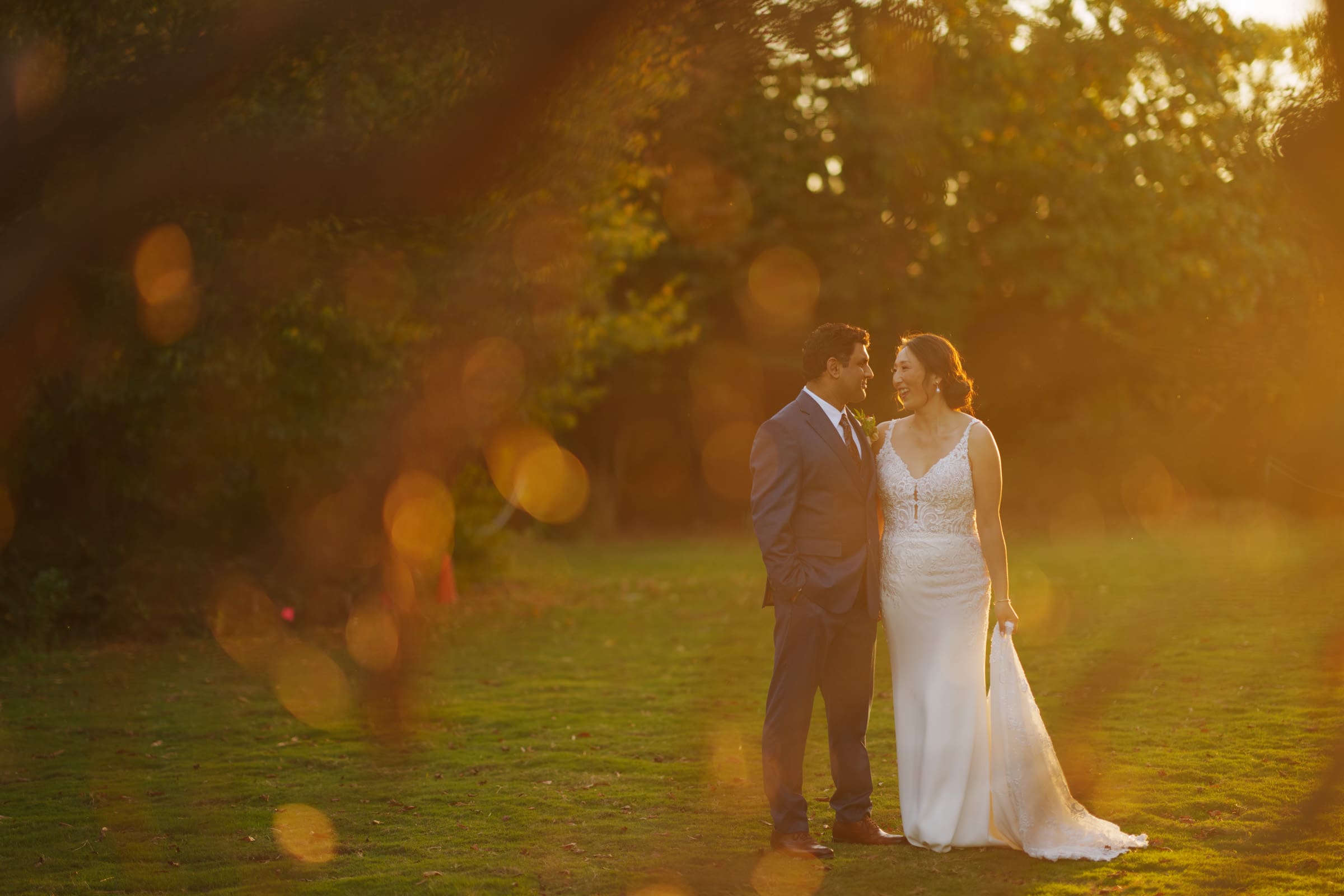 In a sunlit grassy field at the JC Raulston Arboretum, Holly and Sut stand closely together, surrounded by warm, glowing bokeh. The groom wears a suit while the bride is in a white dress, both gazing at each other affectionately.