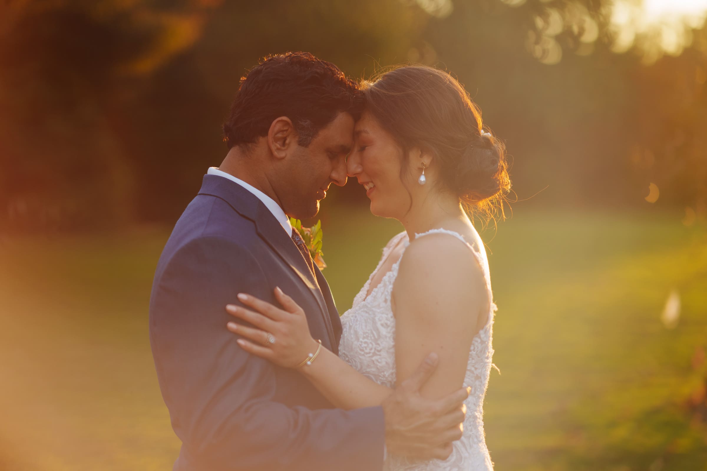 A couple stands closely, gently touching foreheads, bathed in warm, golden sunlight at the JC Raulston Arboretum. The man wears a blue suit, and the woman is in a white lace dress. The sunlit garden provides a blurred backdrop to Holly and Sut's enchanting moment.