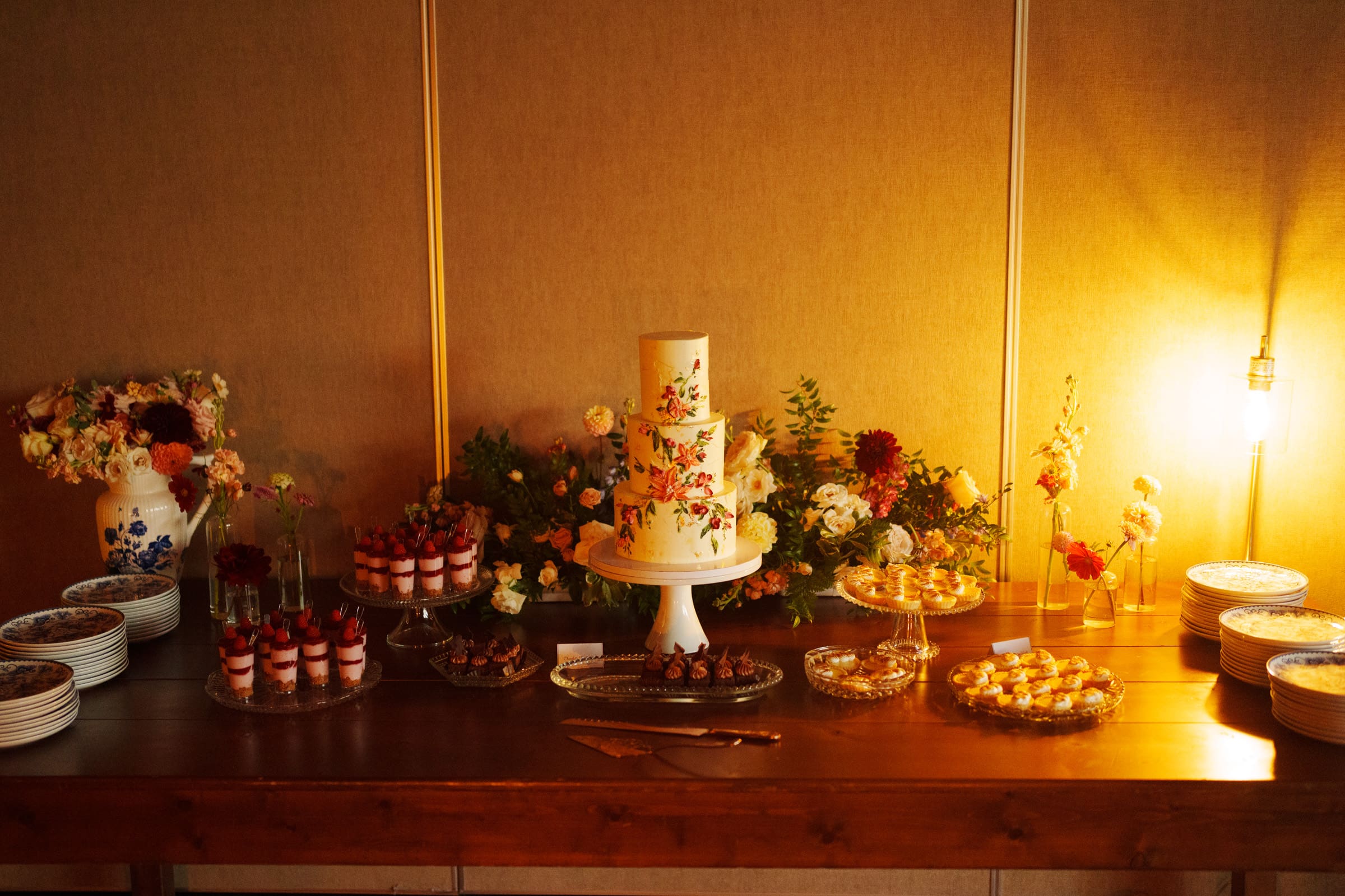 A dessert table at Holly and Sut's JC Raulston Arboretum wedding featured a floral cake centerpiece, surrounded by an array of pastries and sweets. Decorative flowers and stacked plates enhanced the setting, illuminated by warm lighting, creating an inviting and elegant atmosphere.