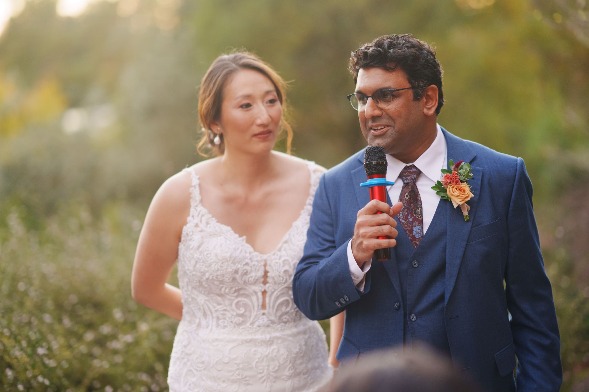 At the JC Raulston Arboretum wedding, Holly in her white lace dress and Sut in a blue suit with a floral boutonniere stand amidst blurred greenery. Sut speaks into a microphone while Holly looks on lovingly, capturing the magic of their special day.