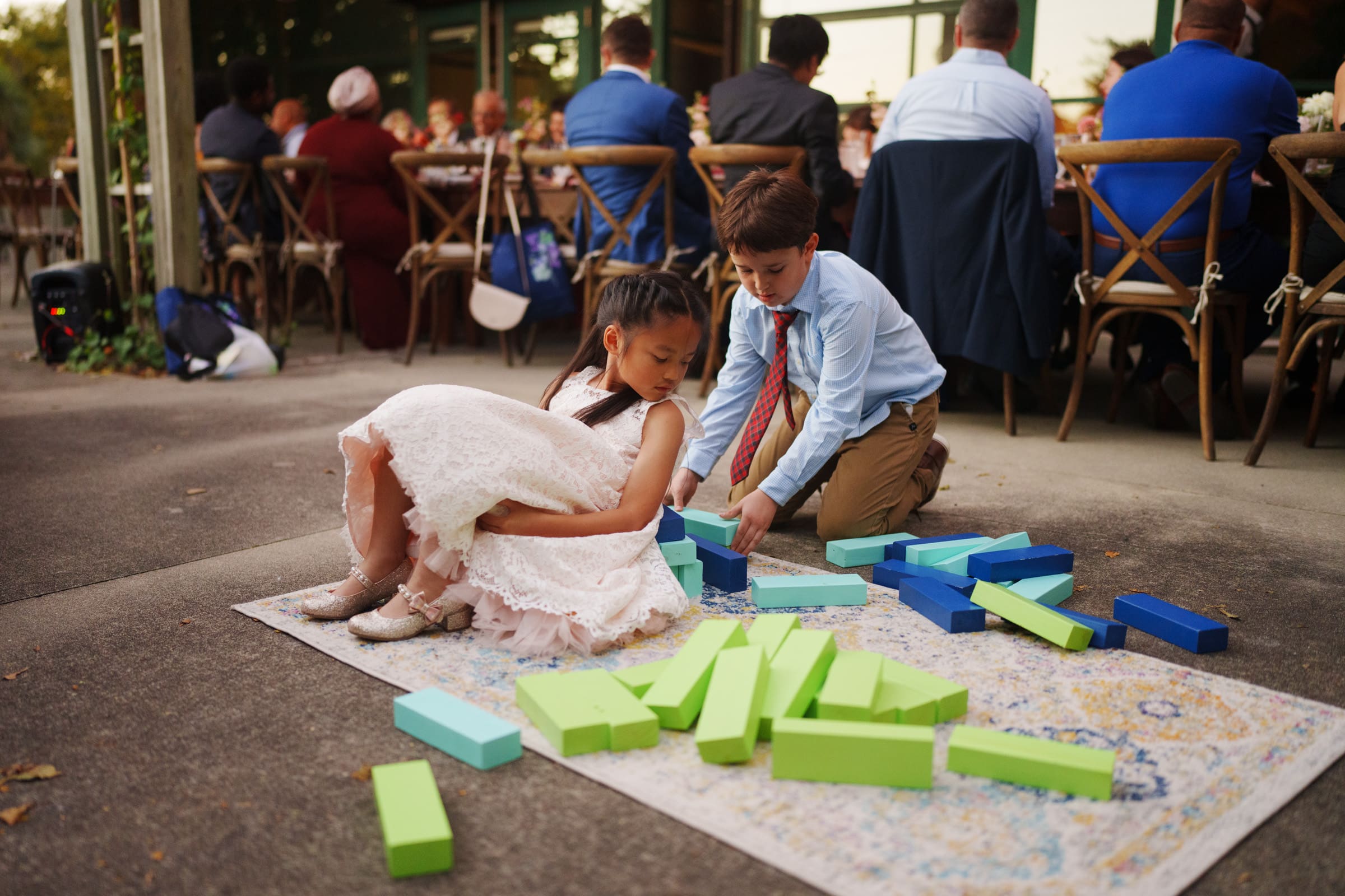 At the JC Raulston Arboretum wedding of Holly and Sut, two children in formal attire joyfully play with large, colorful foam blocks on a patterned rug. A girl in a white dress sits, while a boy in a light blue shirt and tie stacks blocks as adults enjoy the festivities at a nearby table.