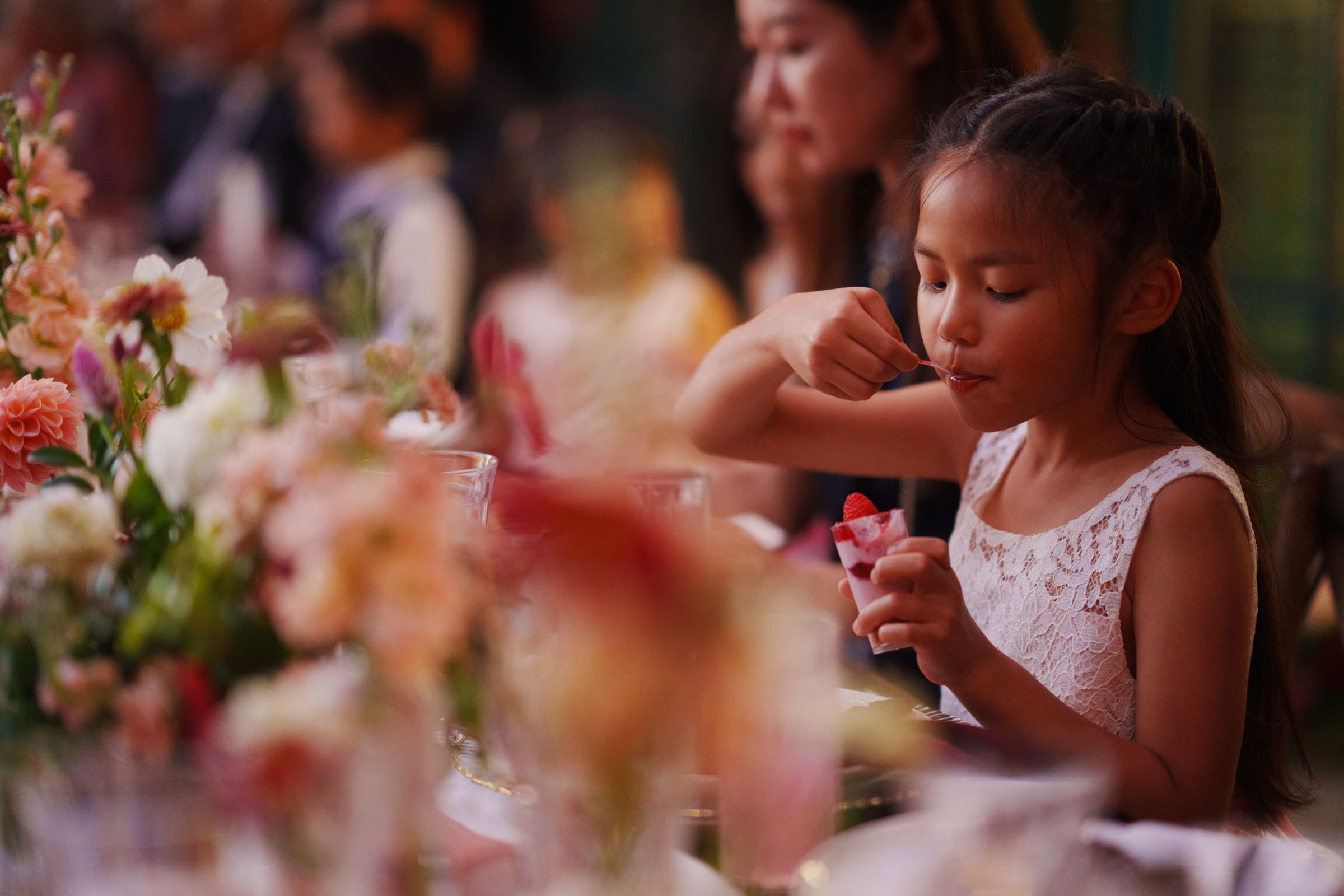 At the JC Raulston Arboretum wedding of Holly and Sut, a young girl in a white dress delights in her dessert amid vibrant flowers and blurred dinner guests. The setting radiates an elegant, joyful atmosphere.