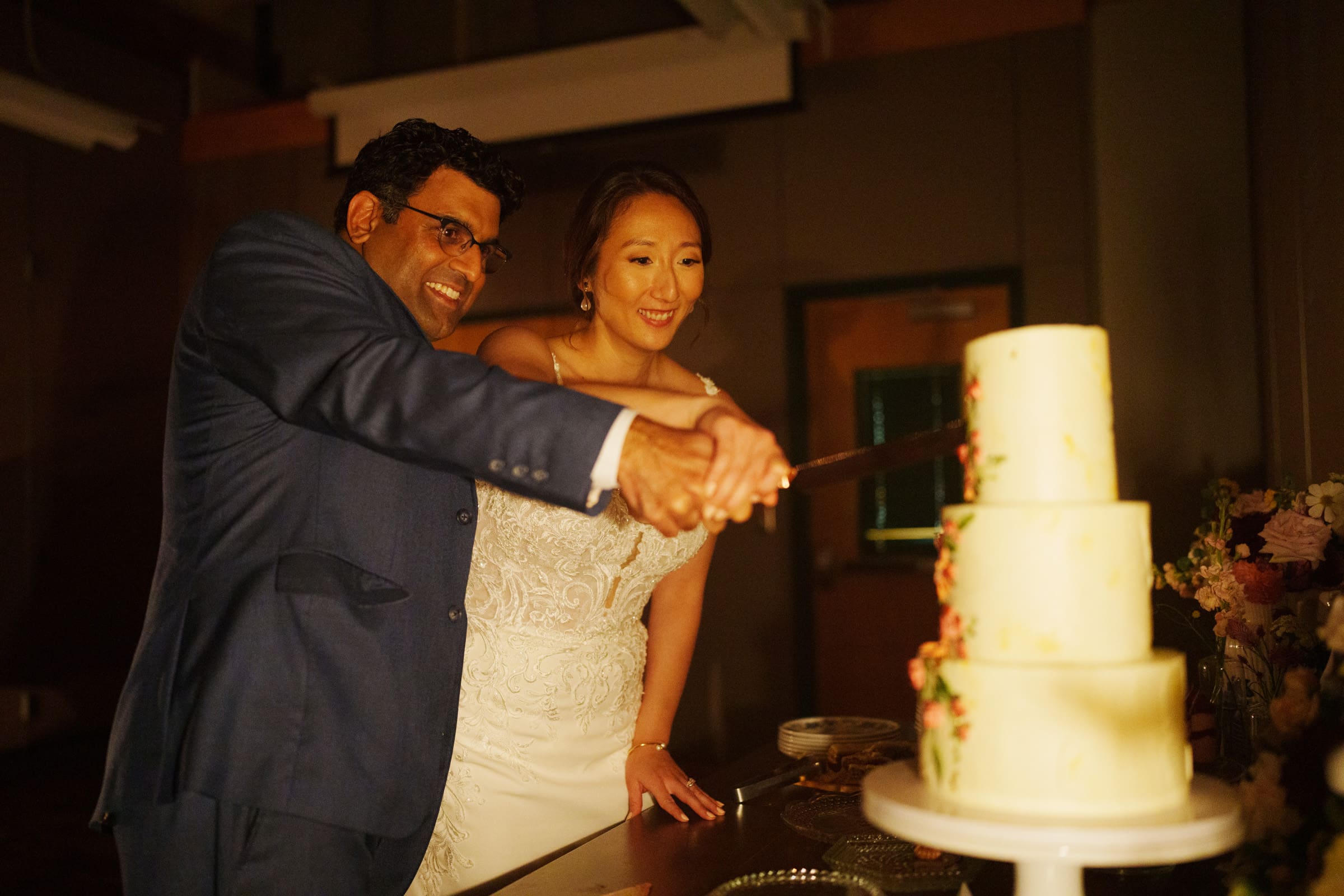 At Holly and Sut's JC Raulston Arboretum wedding, the couple is cutting a three-tiered white cake. The groom is in a blue suit, and the bride wears a stunning white dress. They're smiling warmly as they stand closely together in a beautifully lit room.